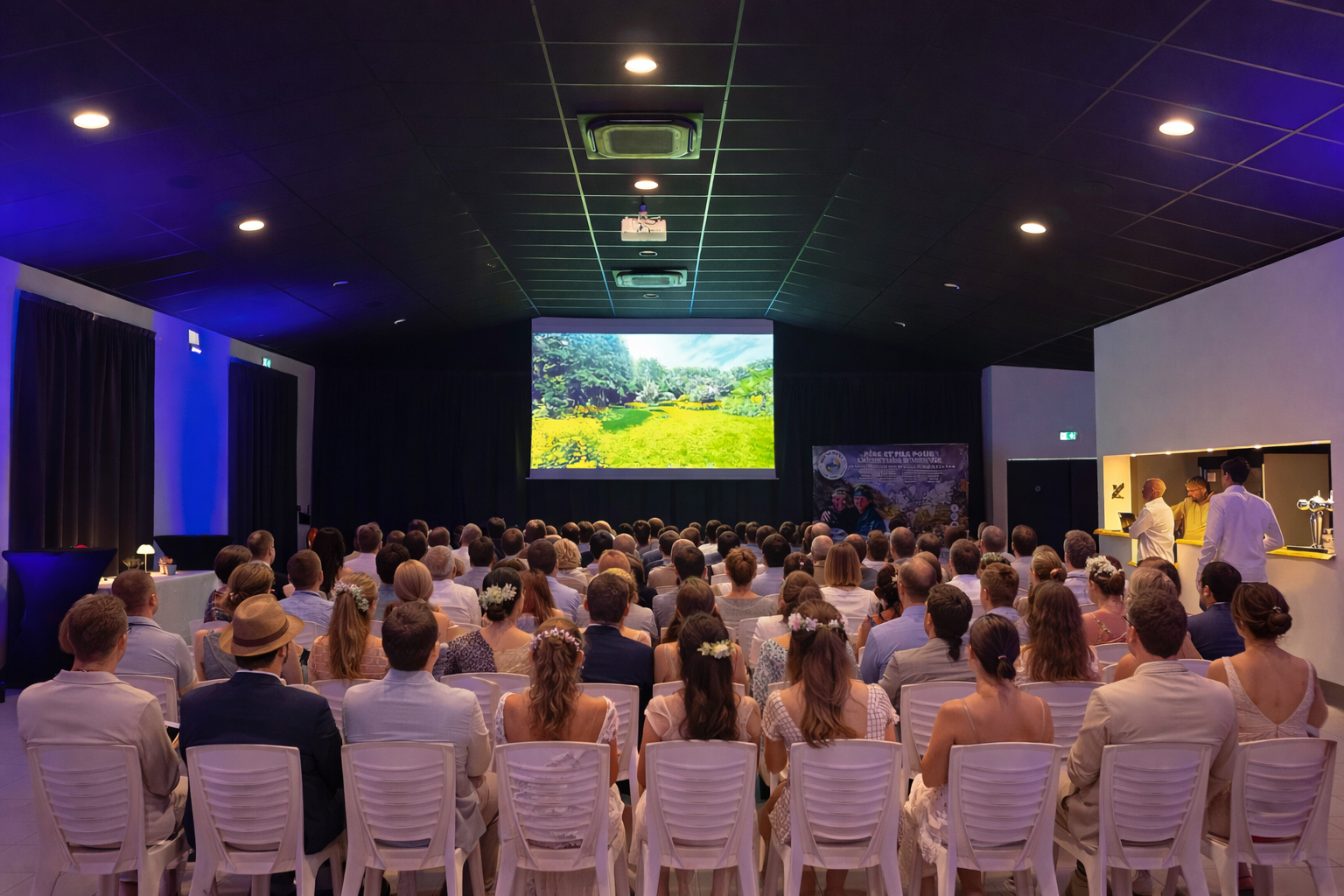 Une salle de projection ou de conférence avec un grand écran affichant une scène de nature verte, remplie de personnes assises en vue de face, regardant vers l'écran. À droite, un comptoir avec des personnes en train de servir ou de discuter.