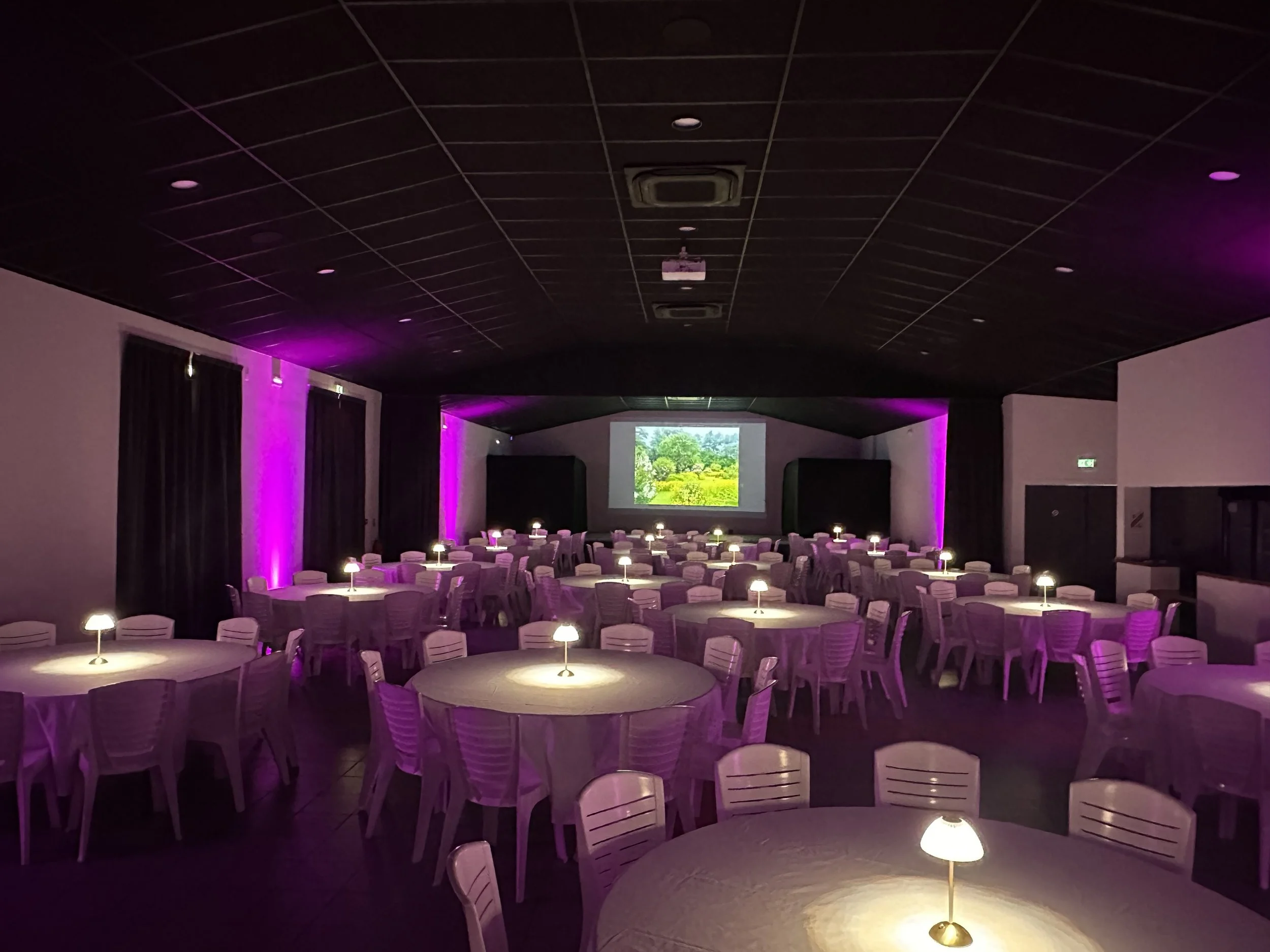 Une salle de réception avec des tables rondes, chaque table a une petite lampe en son centre, des rideaux noirs, deux écran de projection et un éclairage architectural sur les murs.