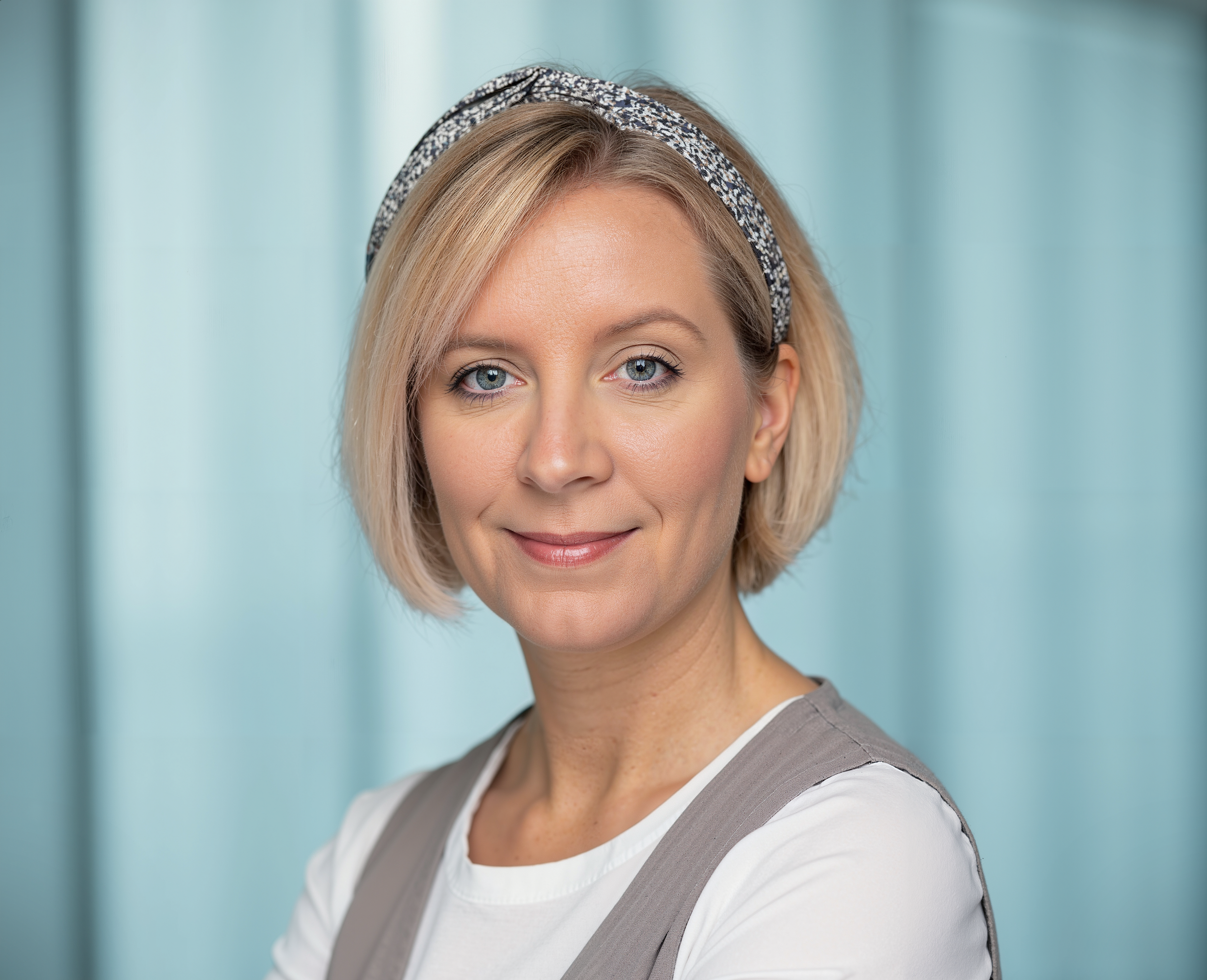 Close-up of a woman with blonde hair, blue eyes, wearing a patterned headband and a gray vest over a white shirt, smiling in front of a light blue background.