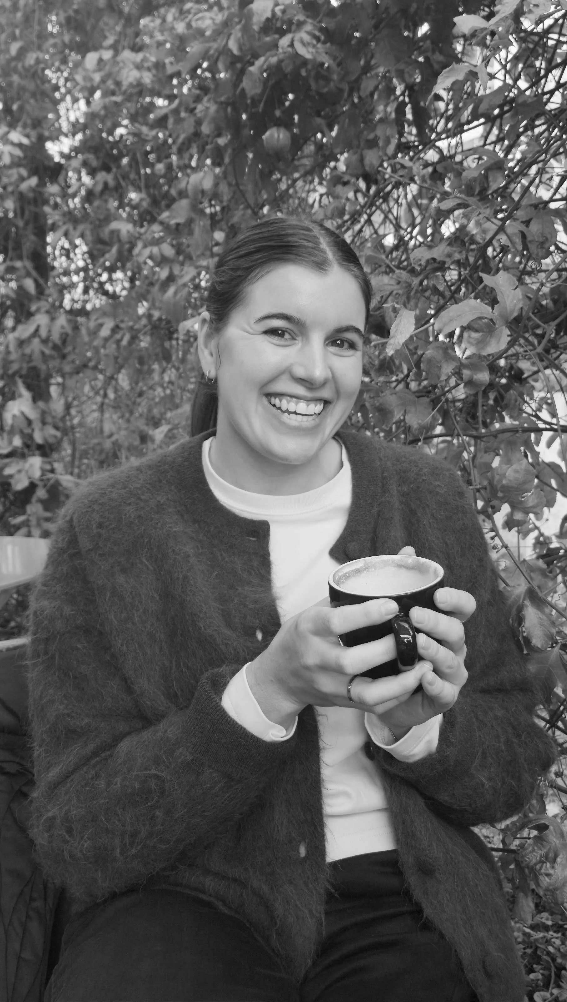 Ira Designs picture smiling and holding a coffee mug outdoors with leafy background.