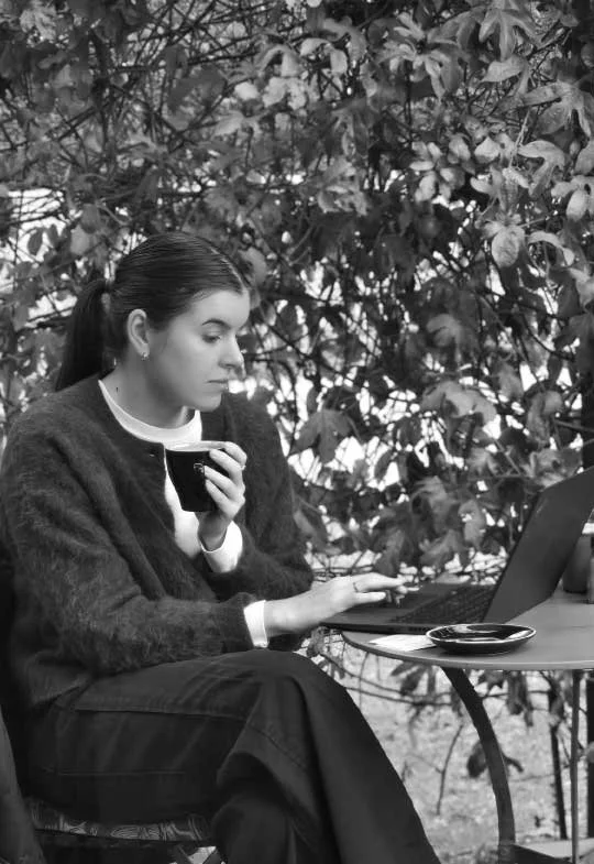 A woman with dark hair tied back, wearing a dark sweater and light shirt, sitting outdoors at a table with a laptop, holding a cup and looking at the laptop screen, with dense leafy foliage in the background.