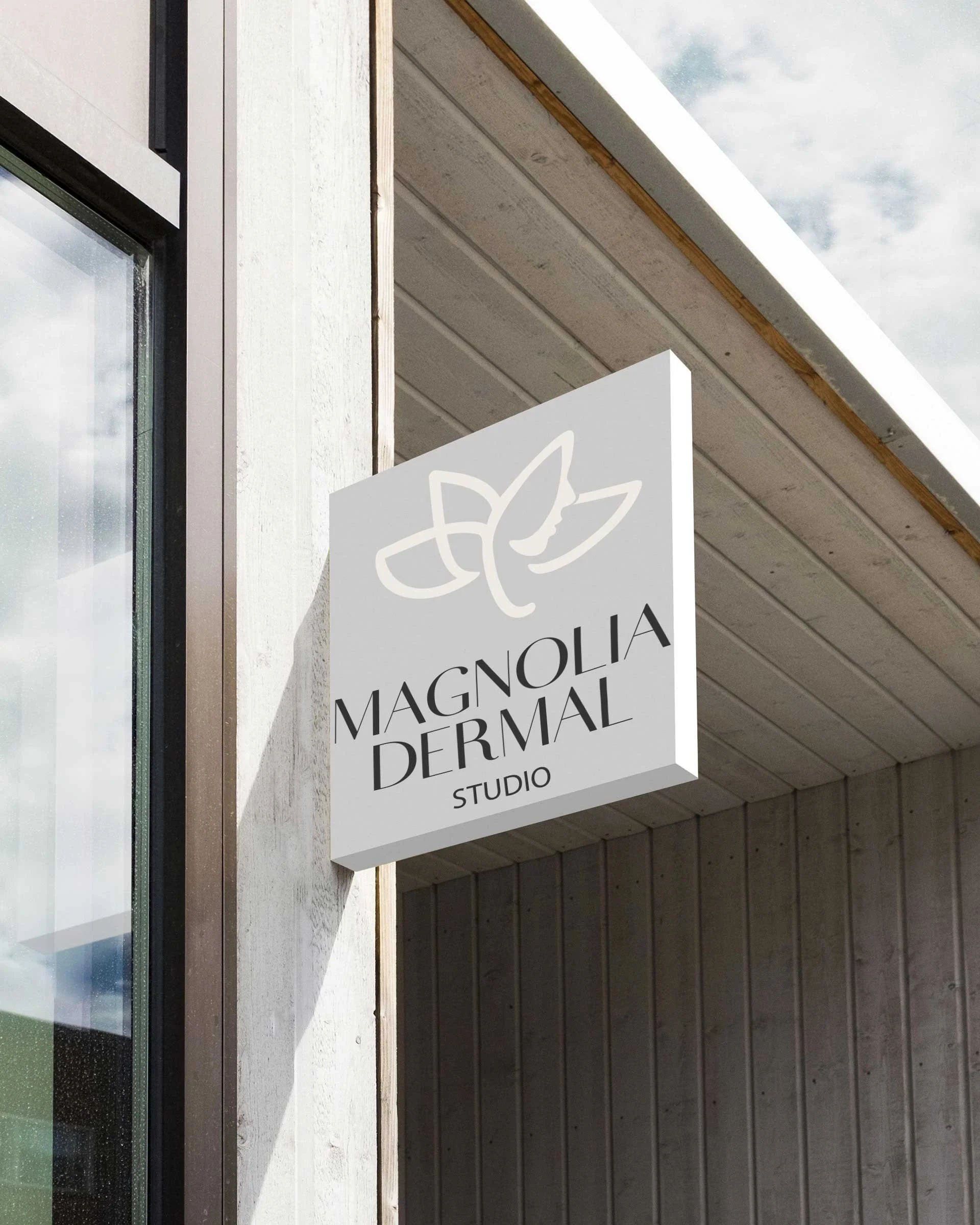 A sign for Magnolia Dermatology Studio hanging outside a building with gray siding, a large window, and white trim.