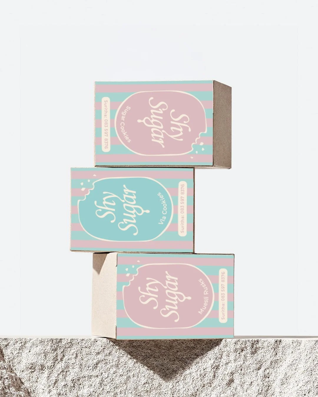 Three small boxes of cookies with pastel pink and blue labels, stacked vertically, placed on a rough white surface with shadows.