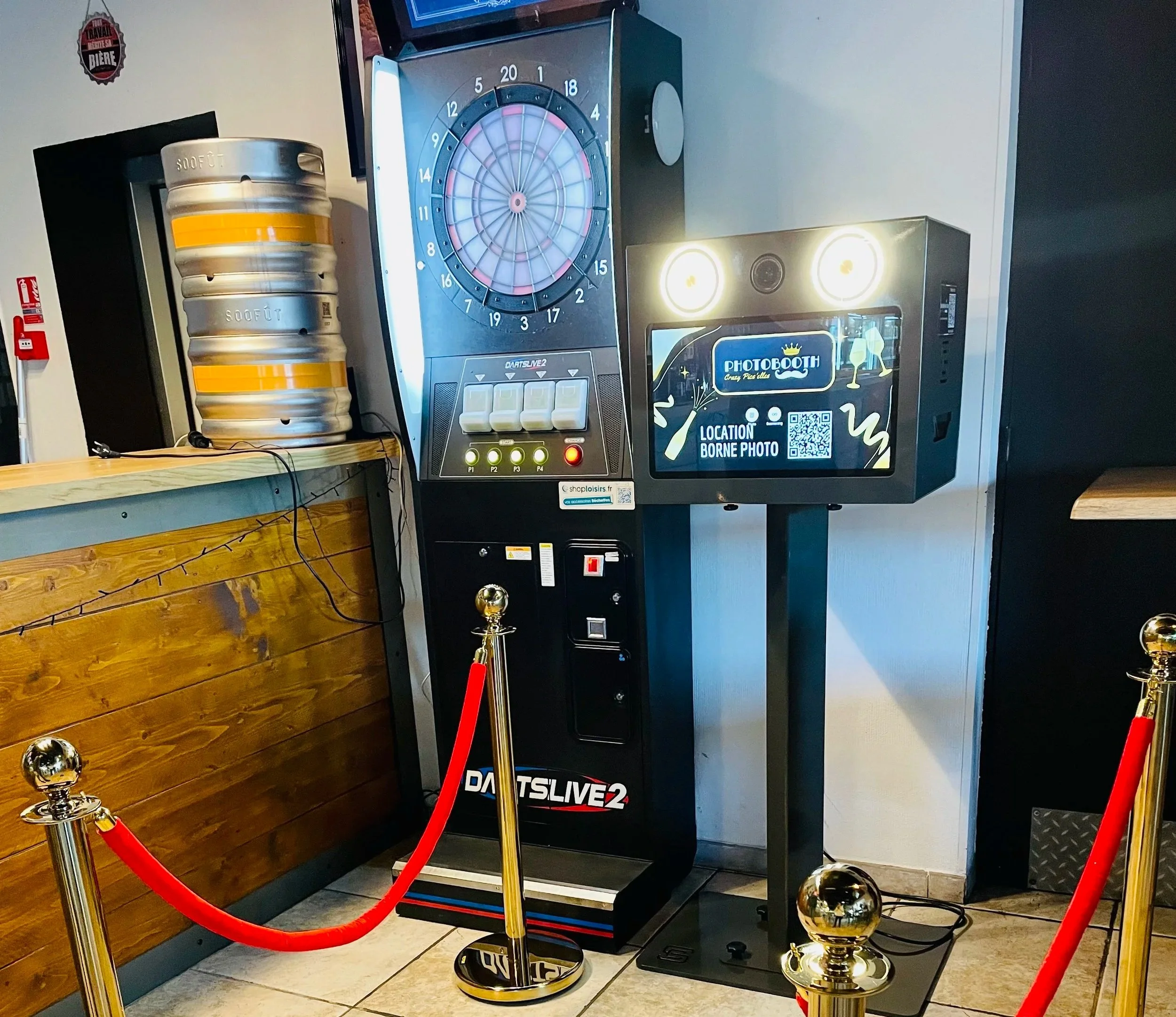 Une machine à jouer au fléchettes électronique, avec un écran pour site de location de bornes photo et un flipper, entourée d'une barrière rouge avec des piquets en métal.