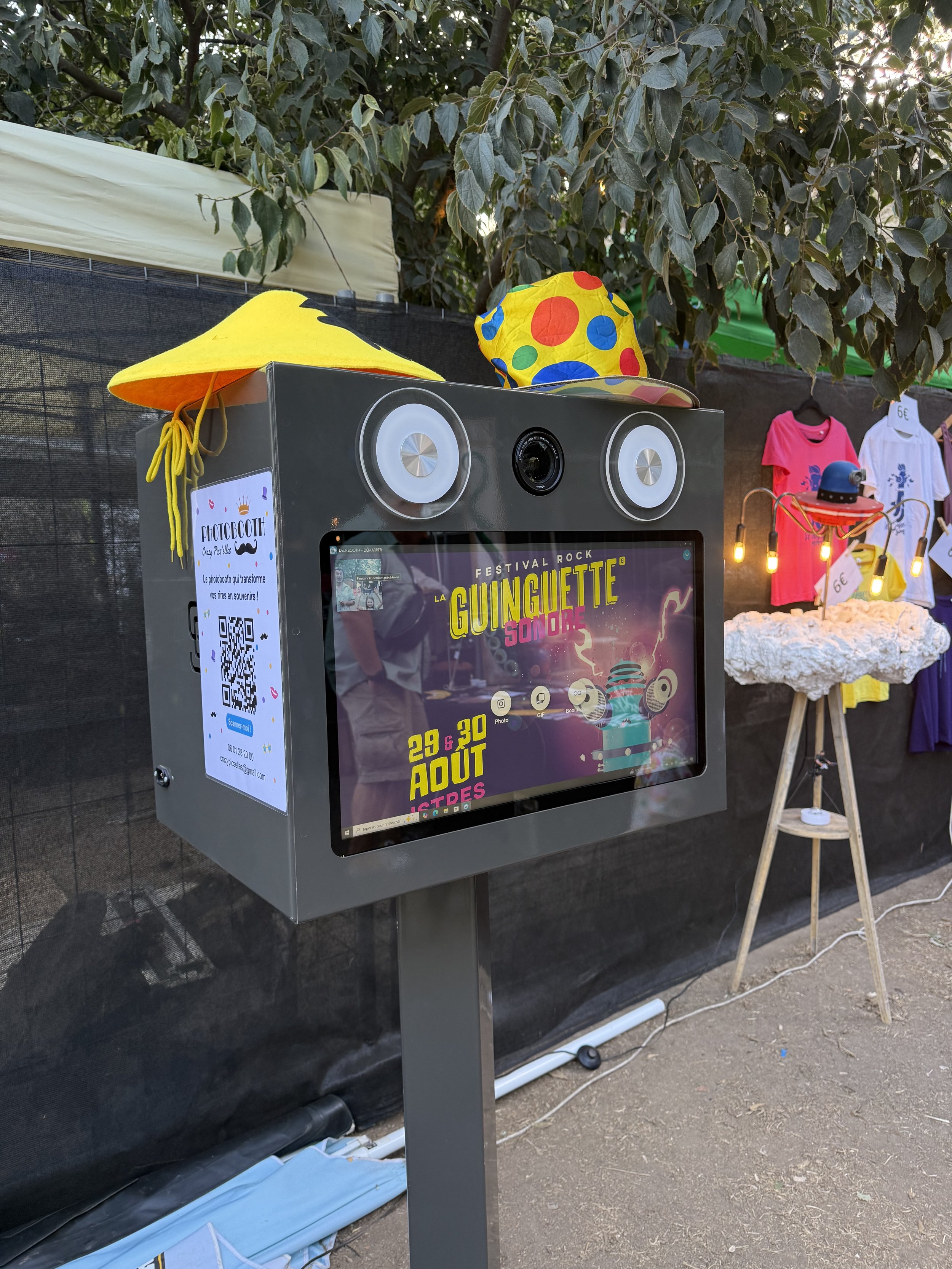 Un photomaton automatisé pour la prise de photos lors du festival rock 'Guinguette Sombre', avec une tablette d'écran, deux haut-parleurs, deux chapeaux lumineux colorés et un décor avec des vêtements pour enfants en arrière-plan.