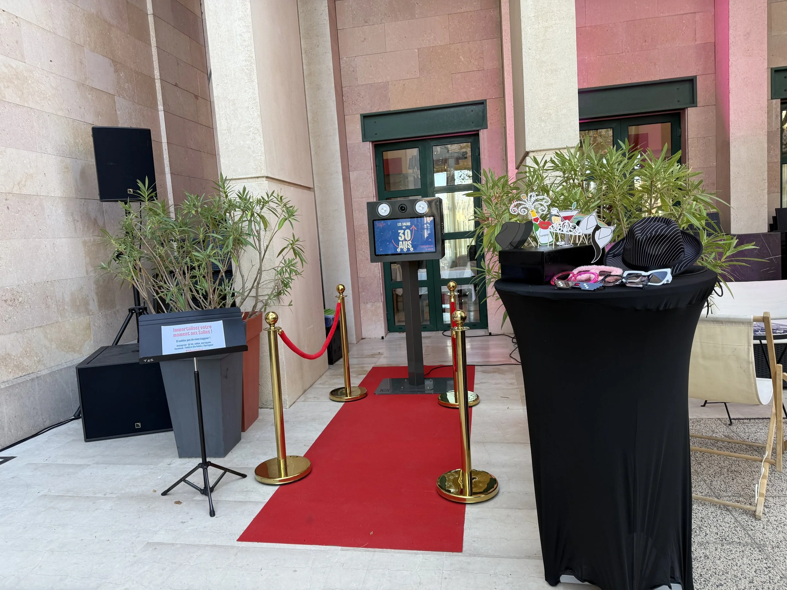 Installation avec un tapis rouge, un panneau avec écriture, une grande plante, une table noire avec des chapeaux, lunettes de soleil et accessoires, un écran digital, et un enceinte à gauche.