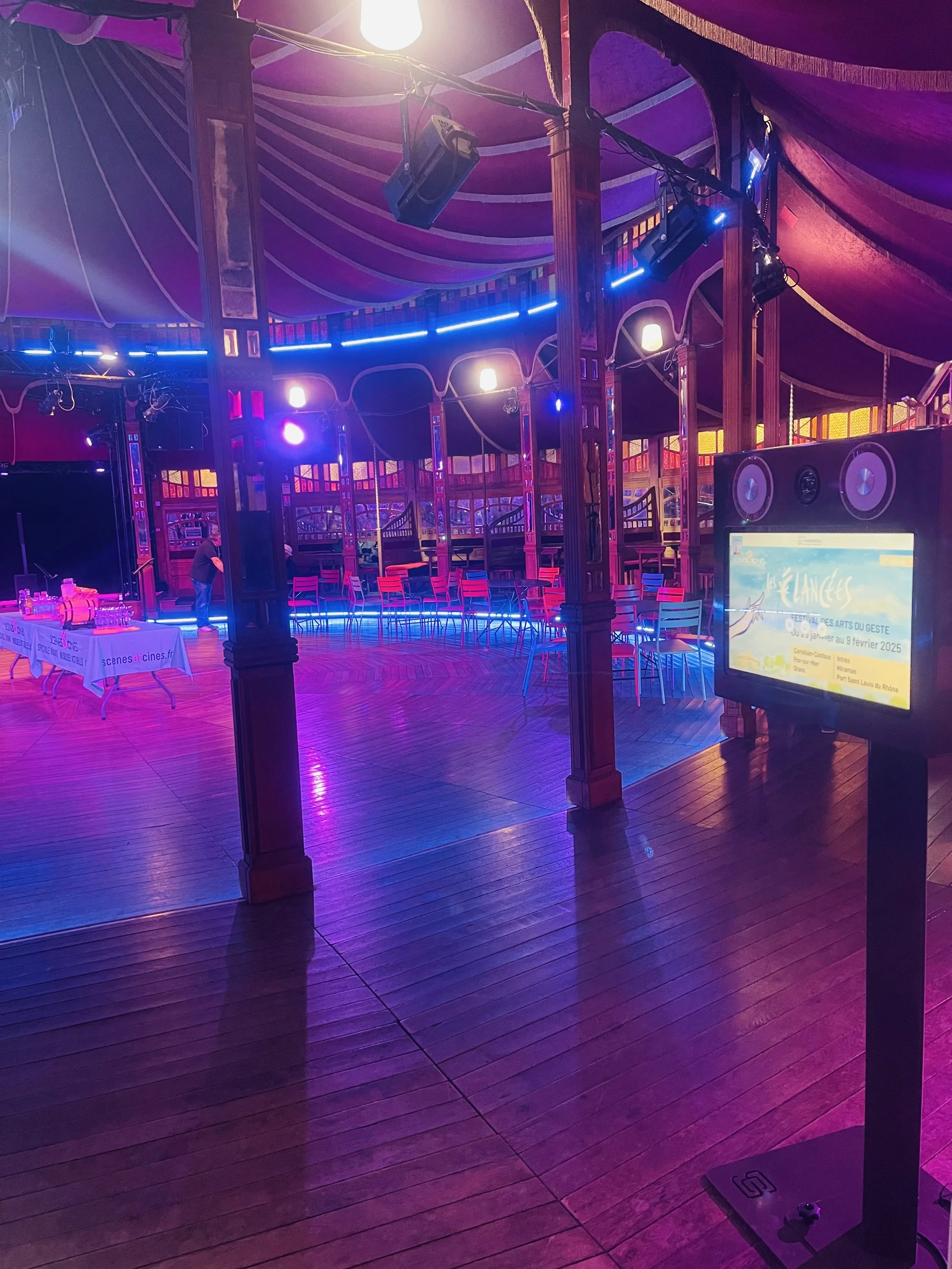 Salle de spectacle ou concert avec lumière colorée, chaises rouges et bleues, scène vide, écran d'information sur le spectacle.