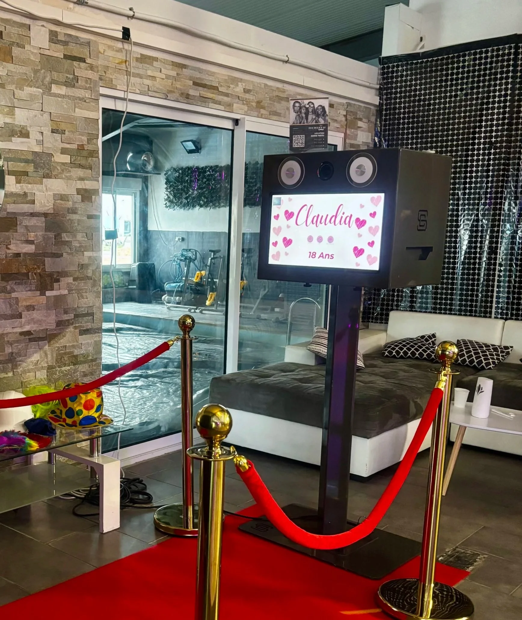 Une borne interactive avec un écran affichant le nom Claudia et la mention 18 ans, entourée de barrières rouges, dans un espace intérieur décoré avec un canapé, des coussins, et une grande baie vitrée.