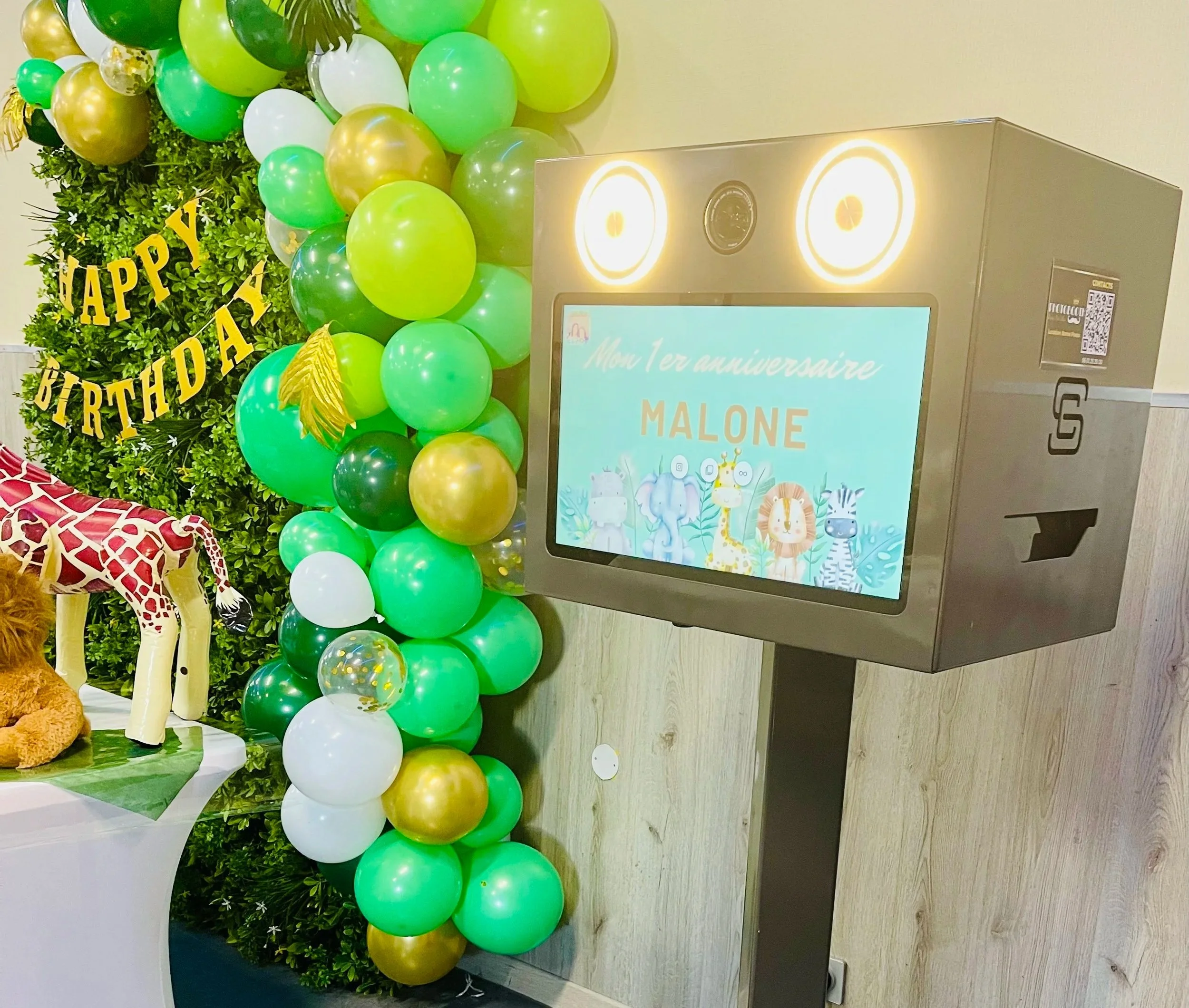 Décoration d'anniversaire pour un enfant avec des ballons verts, or, et blancs, un panneau d'accueil avec le texte "Joyeux anniversaire Malone", un lion en peluche, et un affichage numérique cocciné avec des animaux de la jungle.