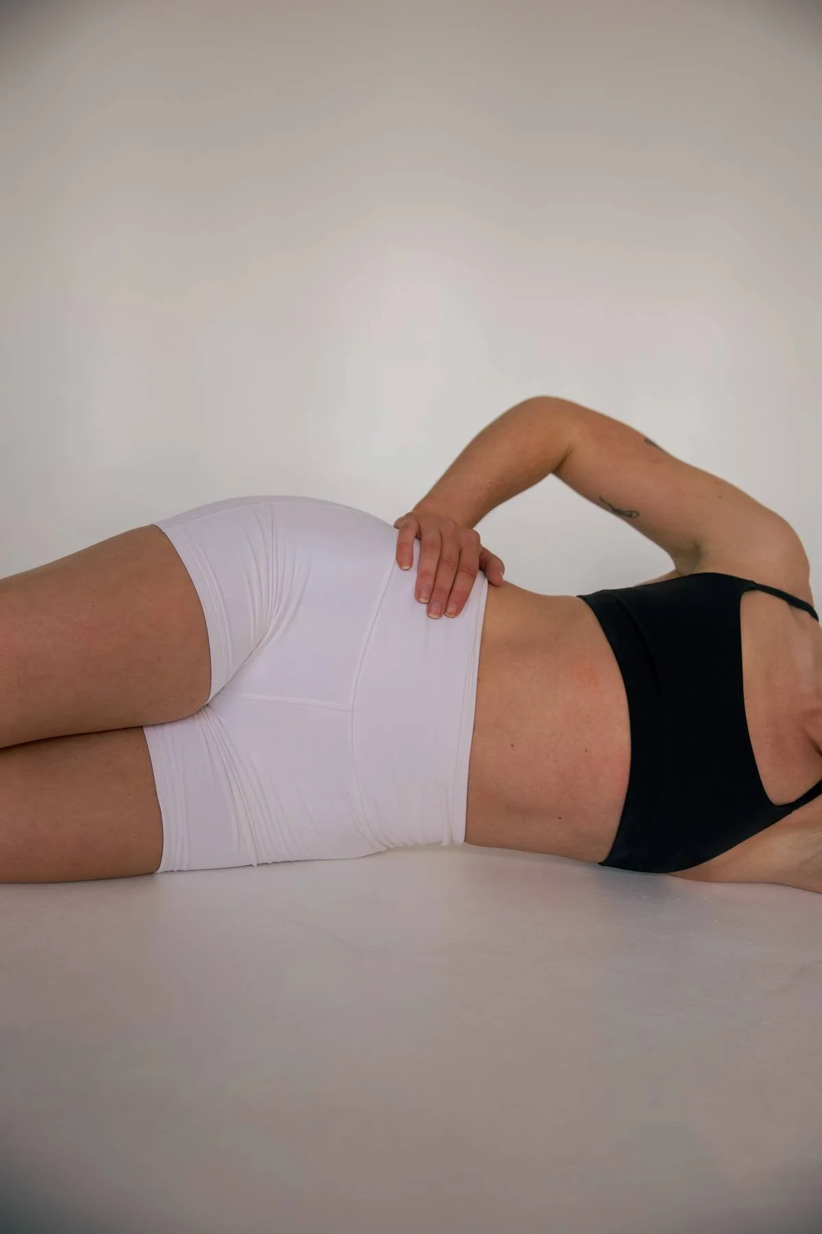A person lying on the floor in a black tank top and white shorts, with one hand resting on their waist and the other supporting their head, against a plain wall background.