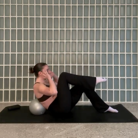 Woman practicing yoga or stretching on a mat with a small exercise ball behind her against a tiled wall.
