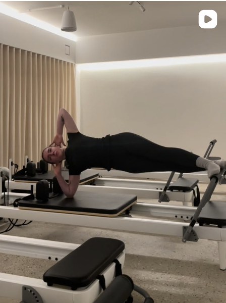 A woman performing a side plank exercise on a Pilates reformer machine in a fitness studio.