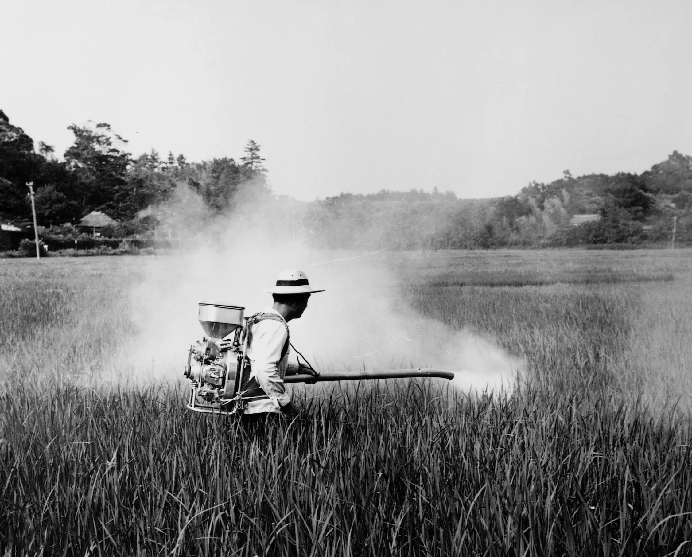 Zgodovinska črno-bela fotografija delavca, ki na polju uporablja zgodnji nahrbtni motorni razpršilnik, kar ponazarja začetke razvoja ECHO in njegovo povezanost s kmetijsko tehnologijo.