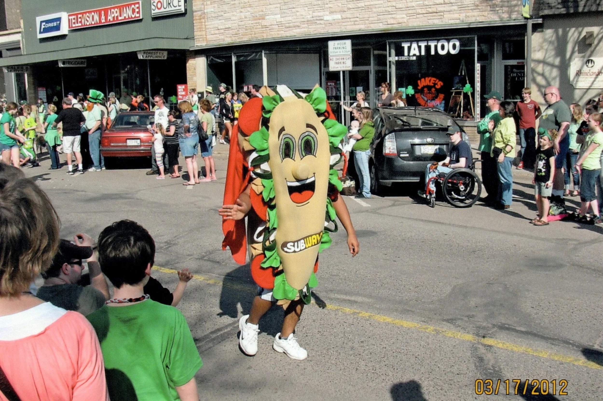 Person dressed in a sub sandwich costume with the Subway logo, walking in a parade on a city street, surrounded by a crowd of spectators.
