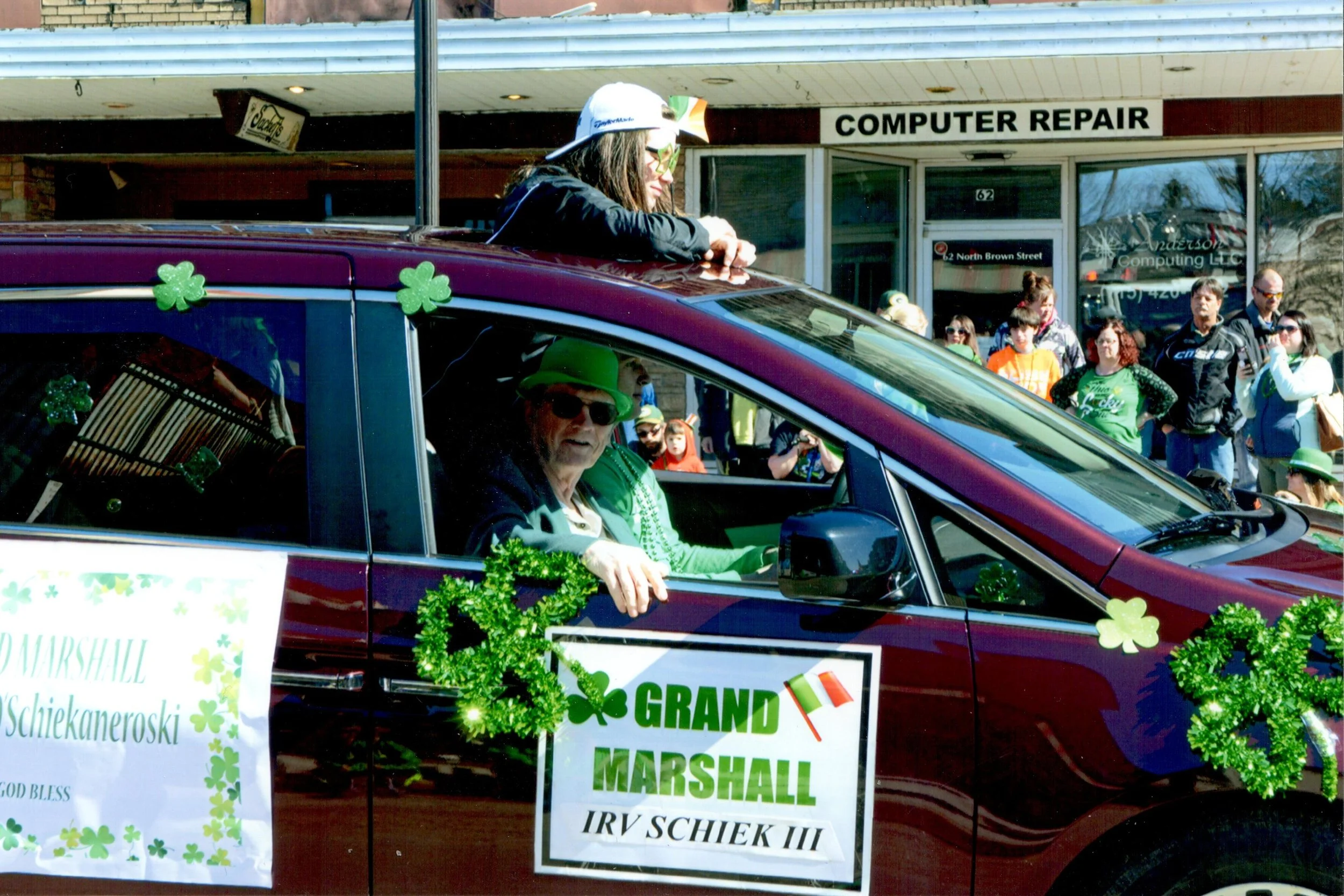 A parade float decorated with shamrocks and green decorations carrying individuals dressed in St. Patrick's Day attire, with a sign that reads 'Grand Marshal IRV SCHIEK III' and St. Patrick's Day symbols, as spectators watch in the background.