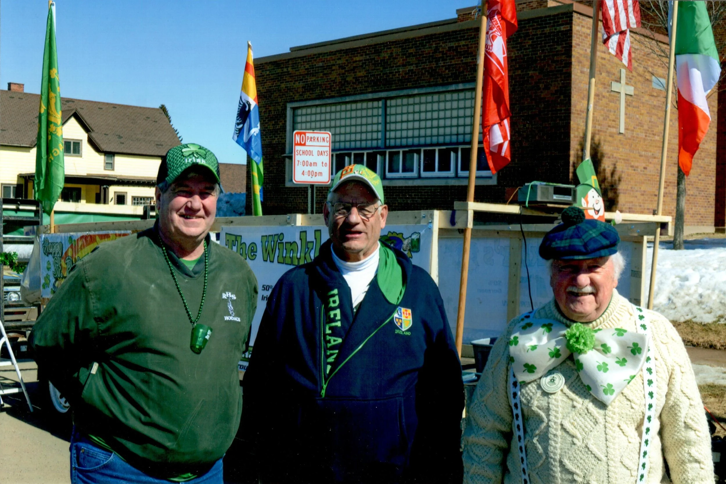 Three men wearing St. Patrick's Day attire standing in front of a decorated float with flags and a cross on a building. One is wearing a green hat and beaded necklace, another a Notre Dame hoodie and cap, and the third a plaid hat and large shamrock 