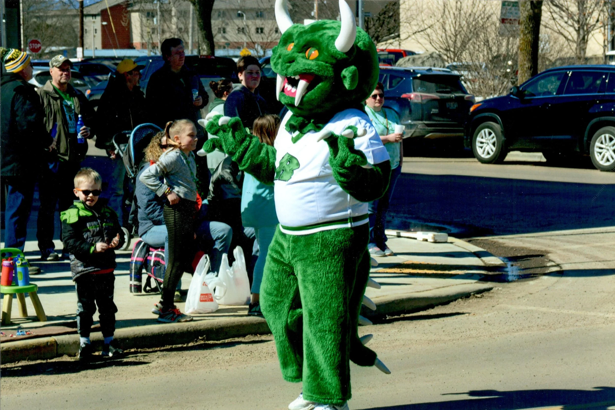 Person in a green dragon mascot costume wearing a white jersey costume with green fur and horns, standing in front of a crowd of children and adults outdoors on a city street during a sunny day.