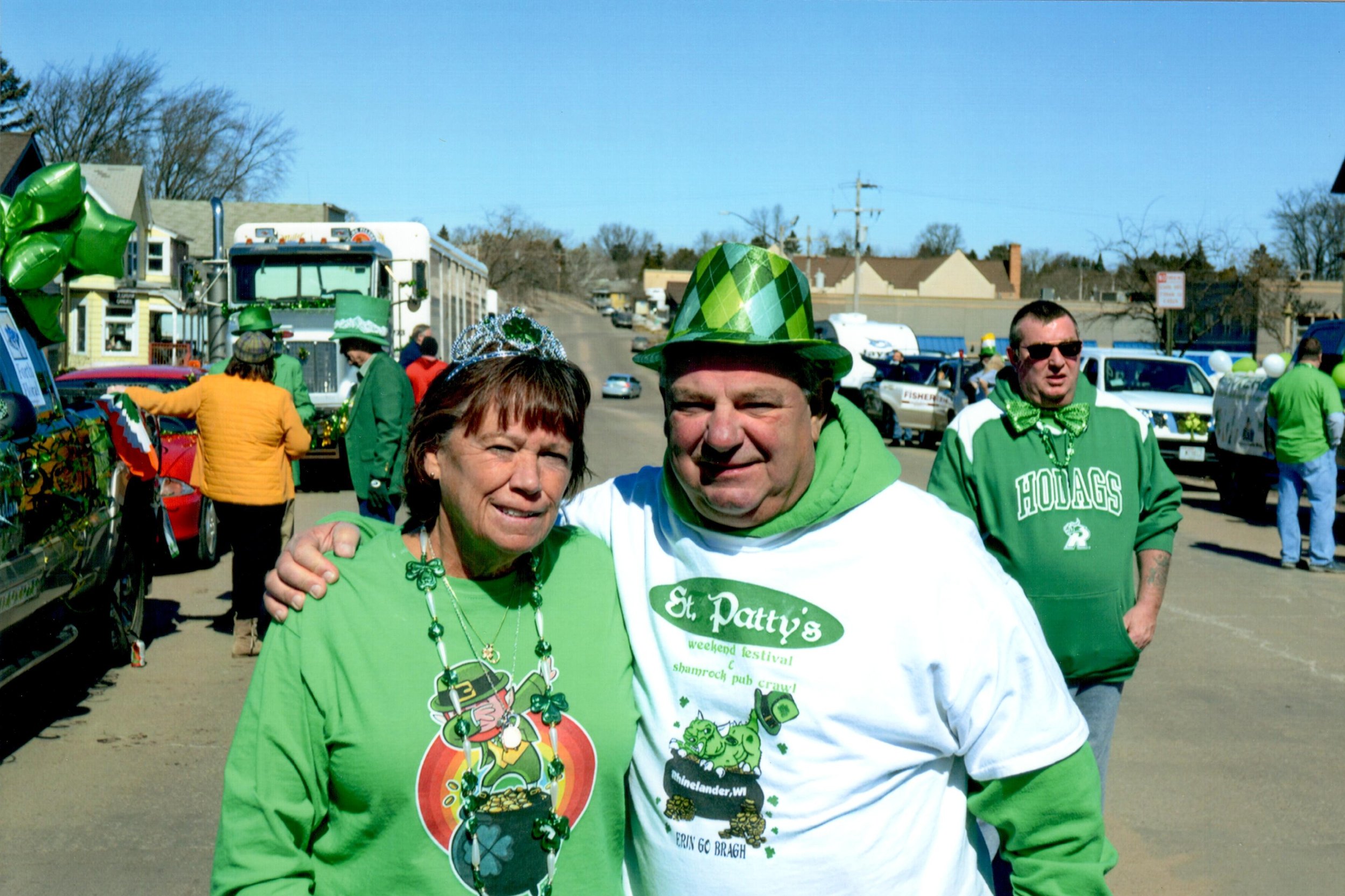 People celebrating St. Patrick's Day outdoors, wearing green clothing and accessories, with balloons and parade floats in the background.