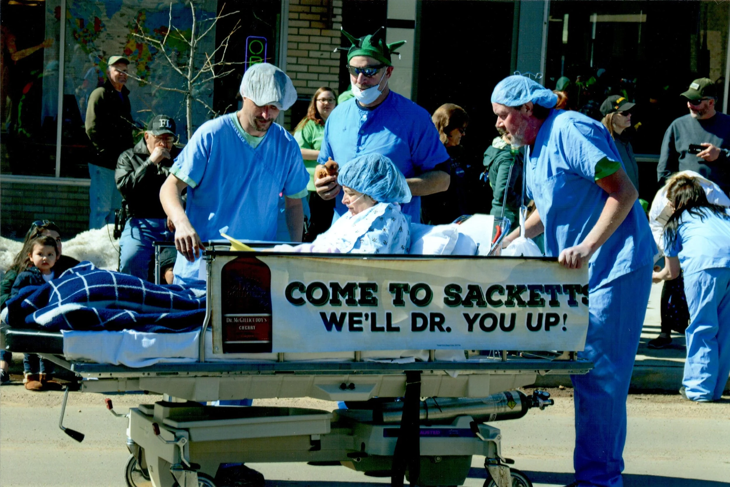 A group of people dressed as surgeons and medical staff in scrubs and surgical caps surrounding a patient in a hospital bed, with a sign that reads "Come to Sackett's, we'll Dr. you up!" on a cart in front of them, during a parade or event.