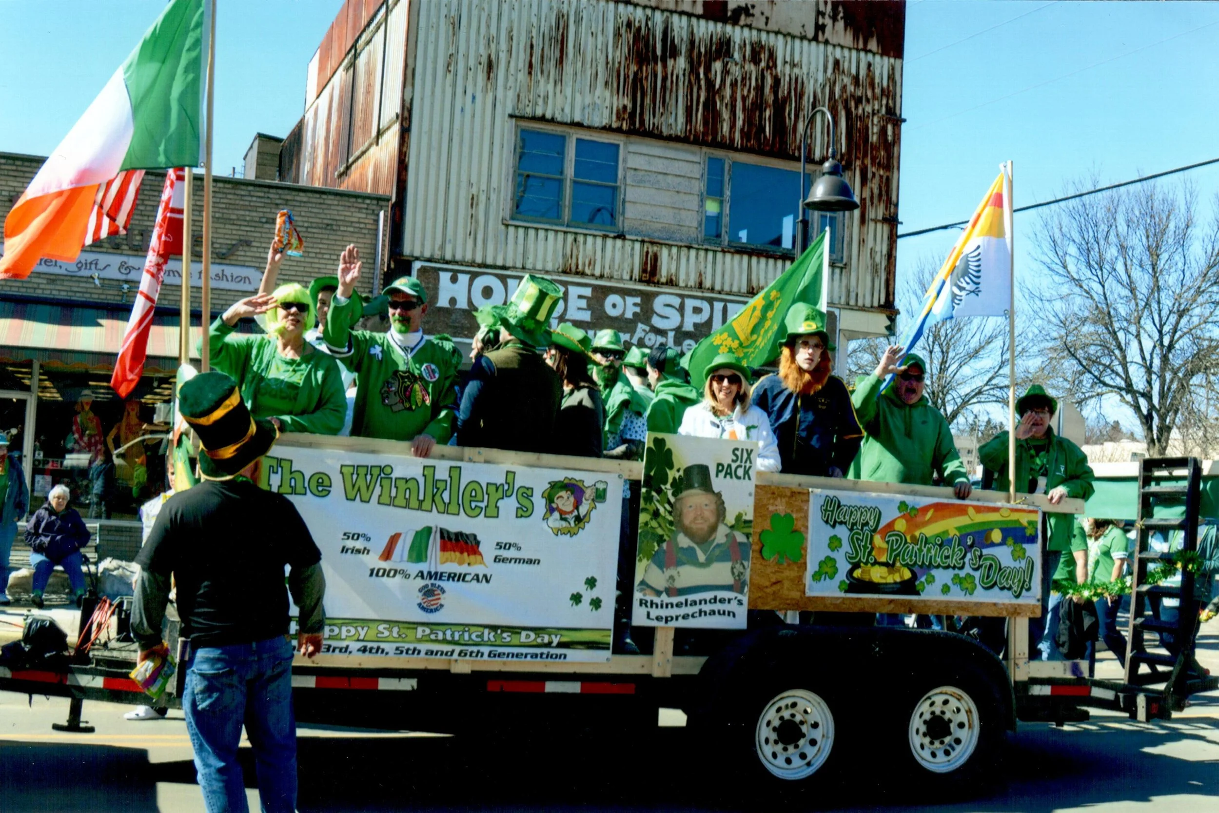 People on a parade float celebrating St. Patrick's Day, decorated with green flags, shamrocks, and festive signs, including one that reads 'Happy St. Patrick's Day' and another with a leprechaun image labeled 'Rhinelander's Leprechaun.'