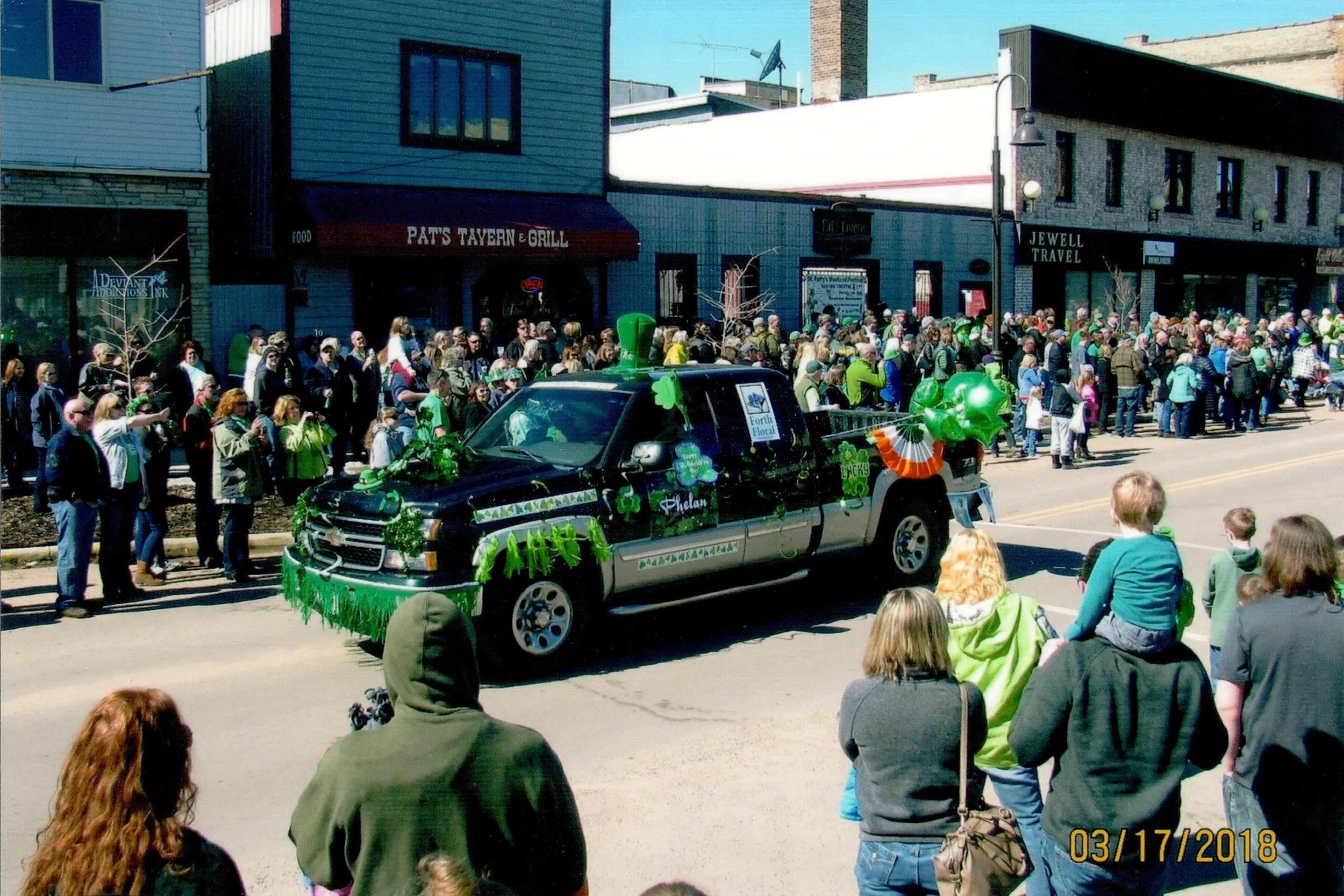 A parade with a decorated black vehicle featuring green shamrocks and Irish accessories, passing through a crowd of spectators on a sunny day.