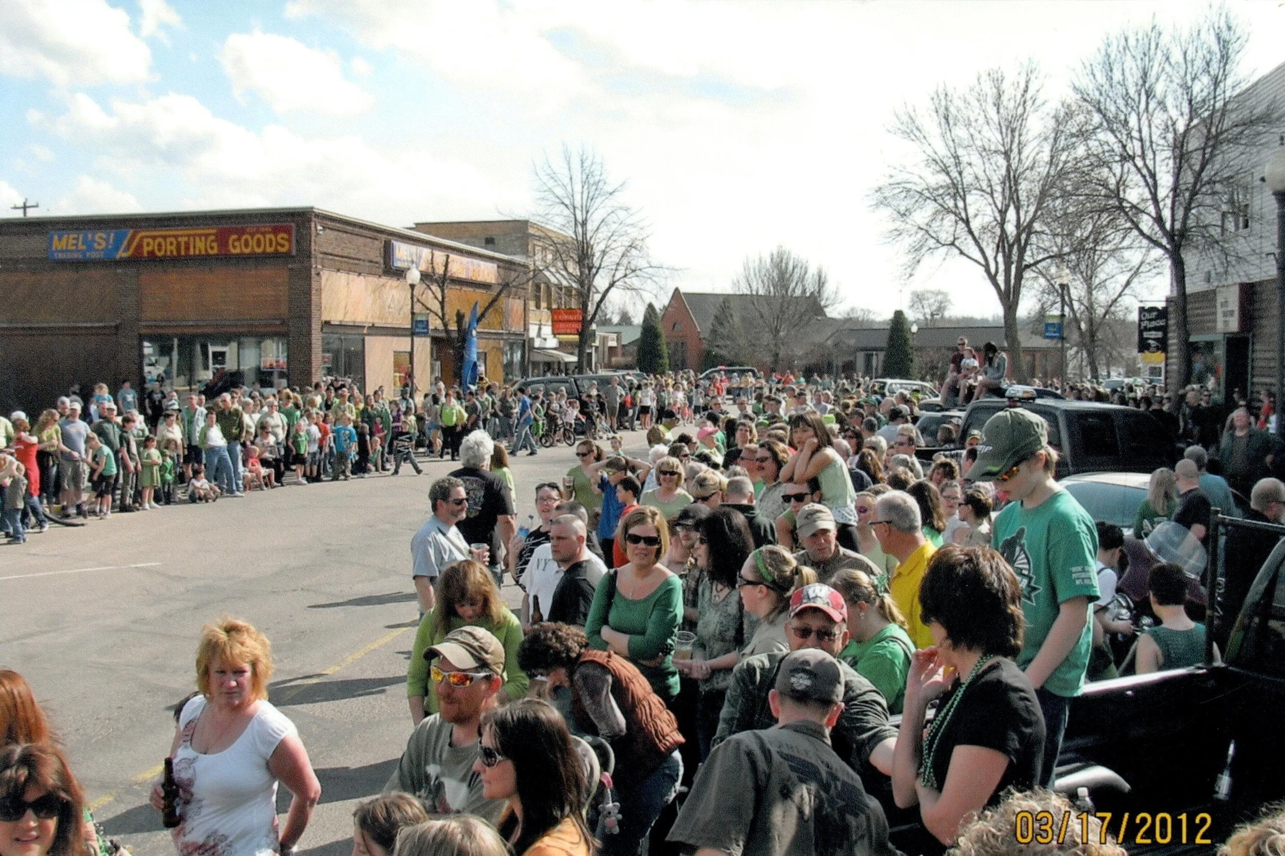 A crowd of people gathered outdoors on a sunny day in front of a store with a sign that reads 'MEL'S PORTING GOODS'. The scene includes various individuals, some sitting, standing, and walking, with clear skies and leafless trees in the background. T