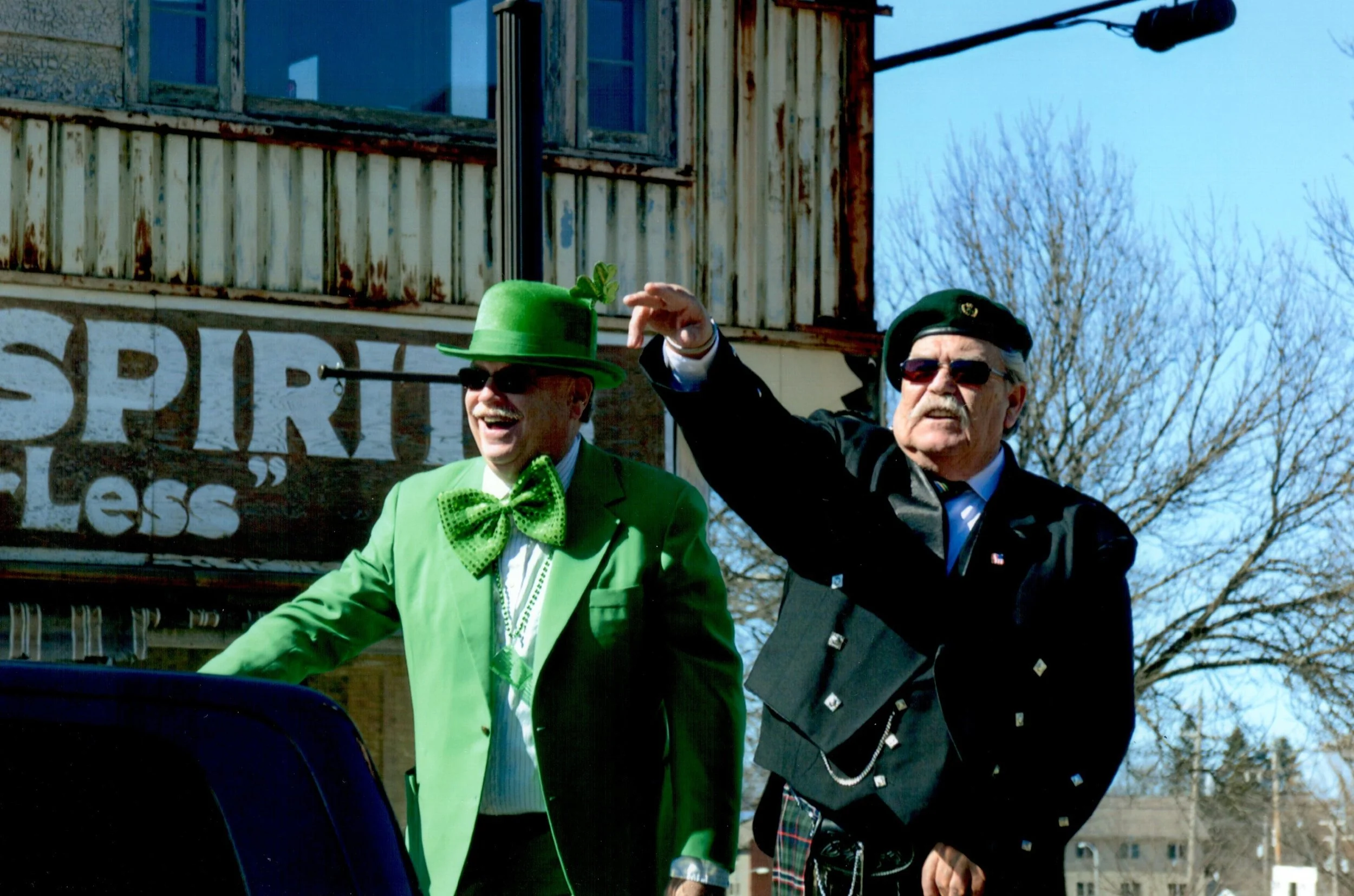 Two men dressed in festive attire, one in a bright green suit with a matching hat, bow tie, and sunglasses, and the other in a black uniform with a beret, sunglasses, and a kilt, waving and smiling outdoors during the daytime.