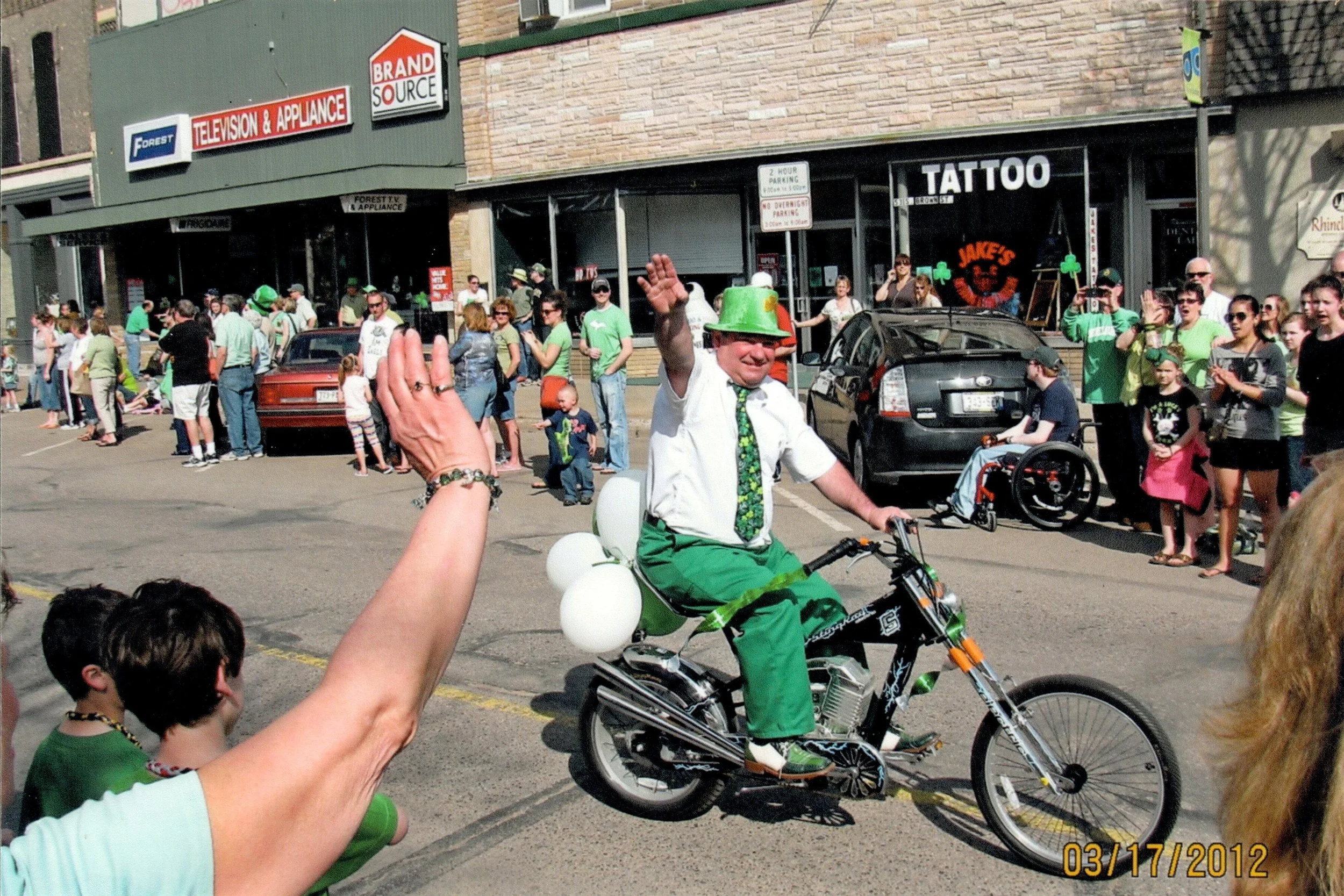 A man dressed in a St. Patrick's Day themed outfit riding a bicycle in a parade, wearing a green hat, tie, and pants, with a crowd of people watching on the street.