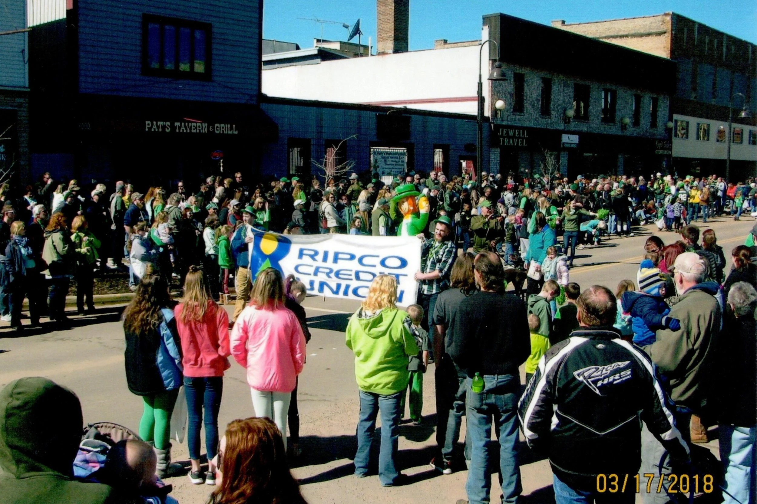 A large crowd of people gathered on a city street, some holding a banner that reads 'RIPCO CREDIT UNION.' The crowd is diverse and appears to be participating in a demonstration or parade. Buildings and shops line the street in the background, and a 