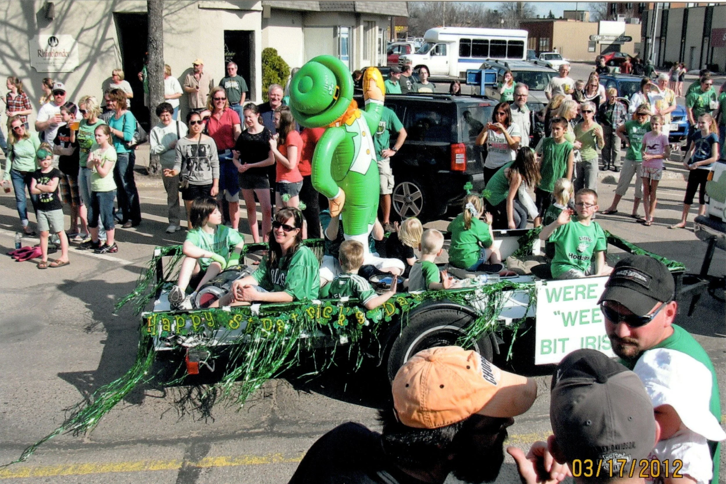 A parade float decorated with a St. Patrick's Day theme, featuring a large leprechaun figure and children wearing green, with onlookers lining the street.