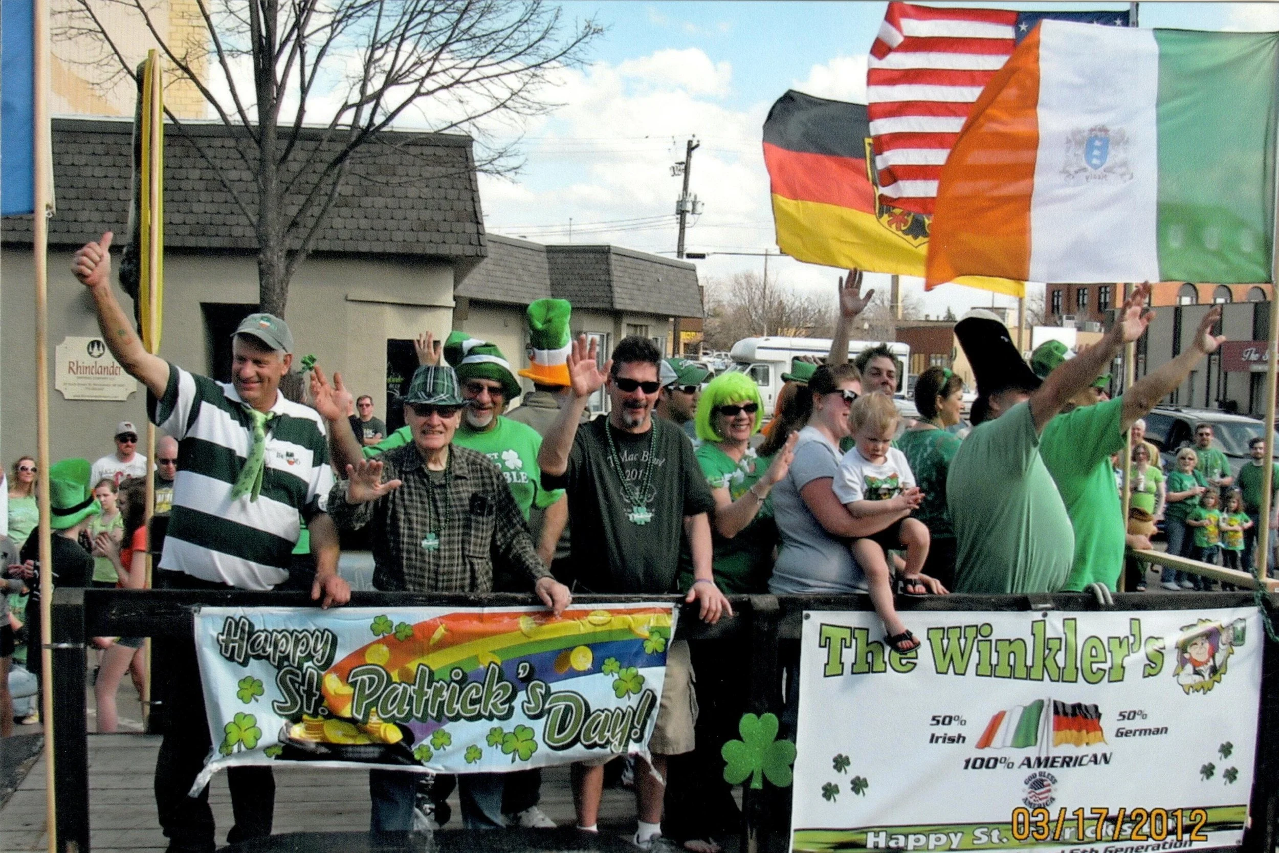 A group of people celebrating St. Patrick's Day parade, wearing green clothing and accessories, waving flags including the Irish, American, Germany, and another flag, with banners reading 'Happy St. Patrick's Day!' and 'The Winkler's'. Some individua
