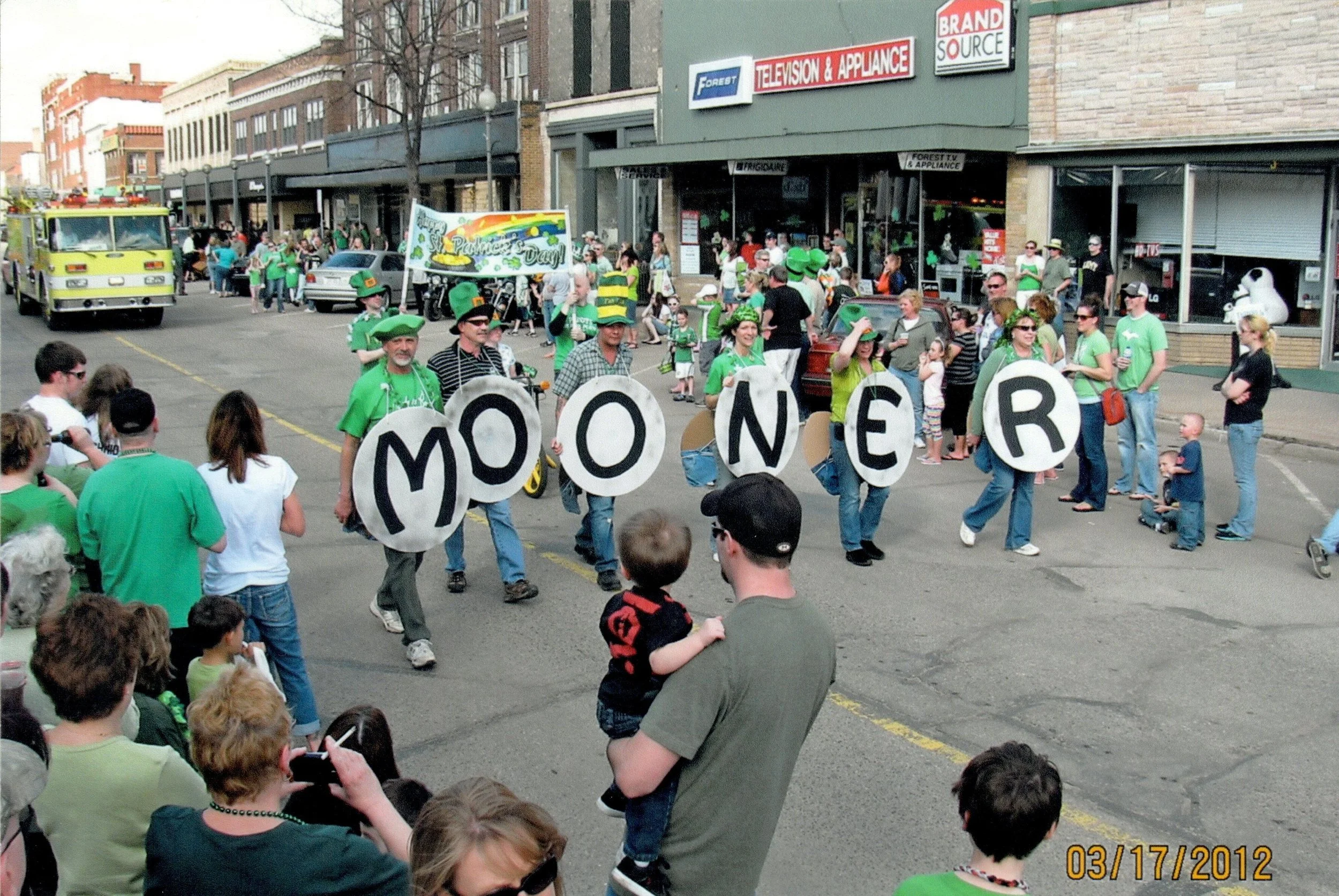 St. Patrick's Day parade with people dressed in green, a sign reading 'MOONER,' and an audience watching on a city street.