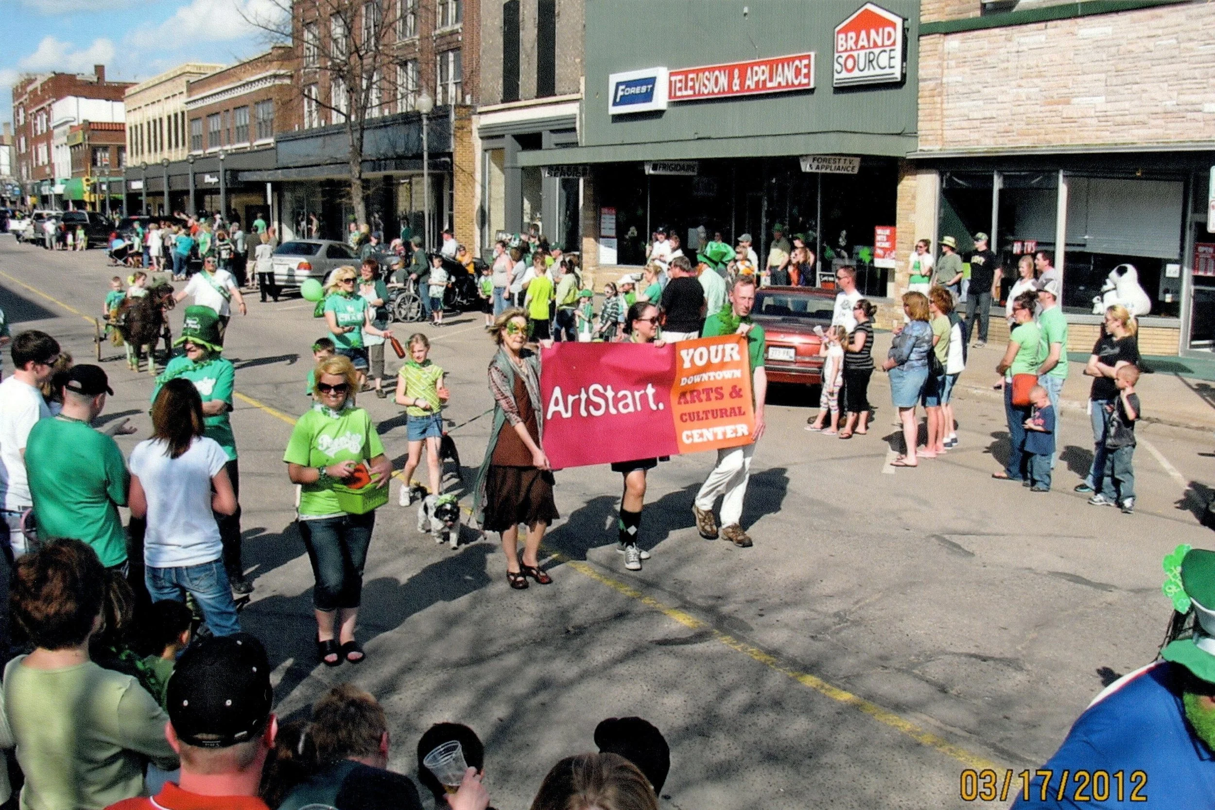 A parade on a downtown street with people gathered along the sidewalk, some holding balloons and wearing green, with a group holding a large red banner reading 'ArtStart Your Downtown Arts & Cultural Center'.