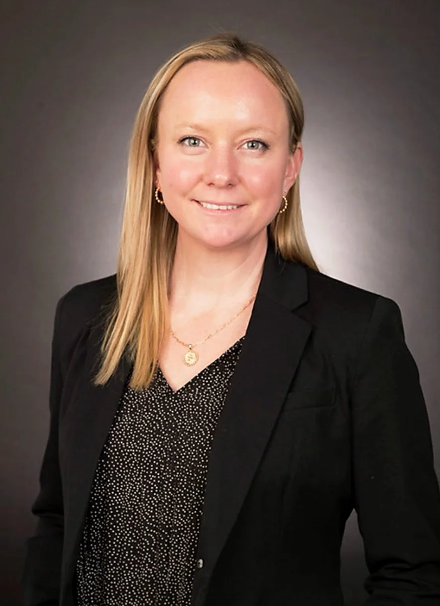 A woman with blonde hair wearing a black blazer, a patterned black and white blouse, gold earrings, and a gold necklace, smiling against a gray background.