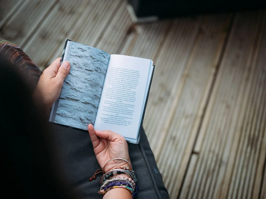 Person holding an open book with textured wall image on one page, and text on the other, sitting on a wooden deck.