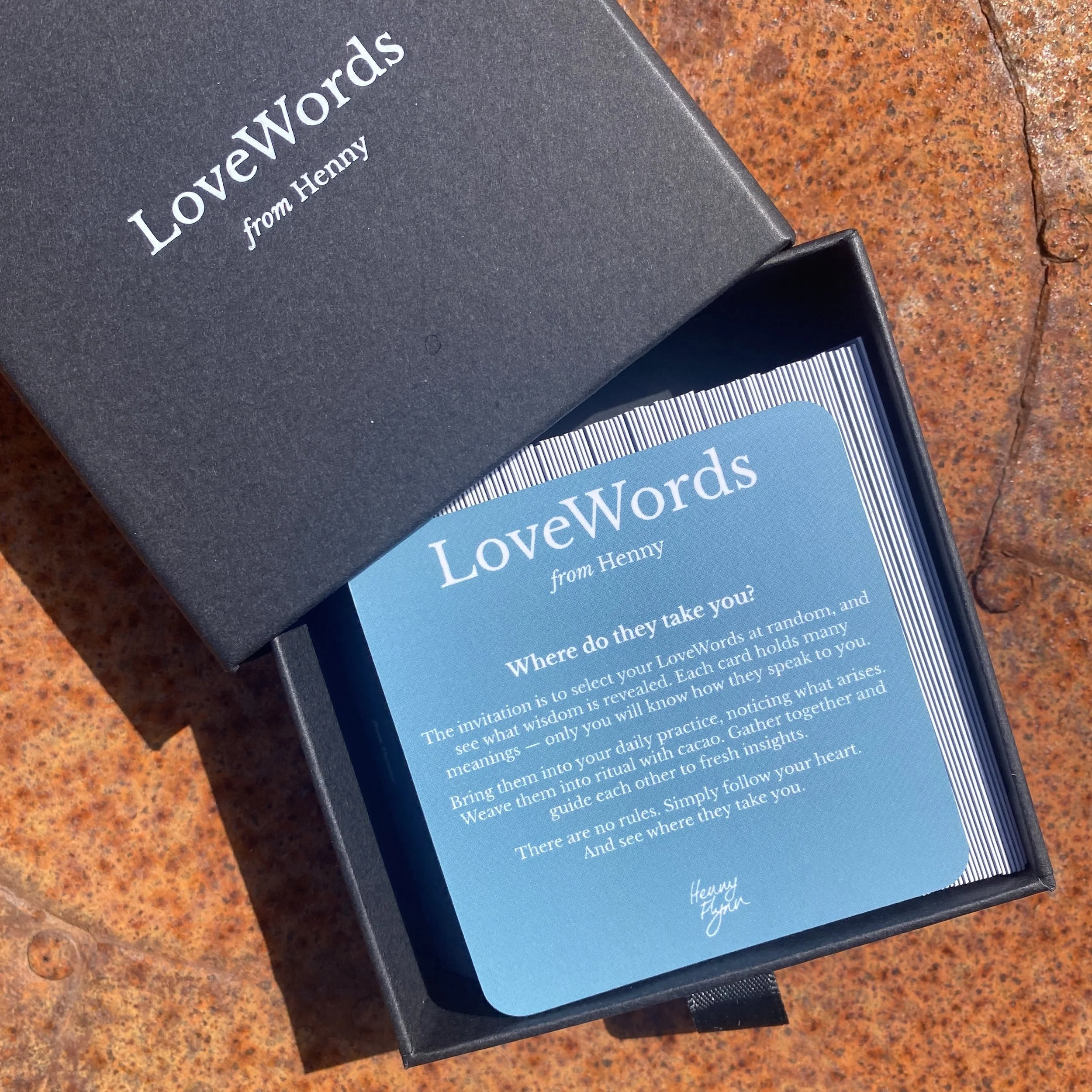 A black box containing a blue card with white text about LoveWords, resting on a rust-colored surface. The card explains the purpose of LoveWords as a guide to understanding the meanings of words through ritual and insight, with a signature from Henry Flynn.