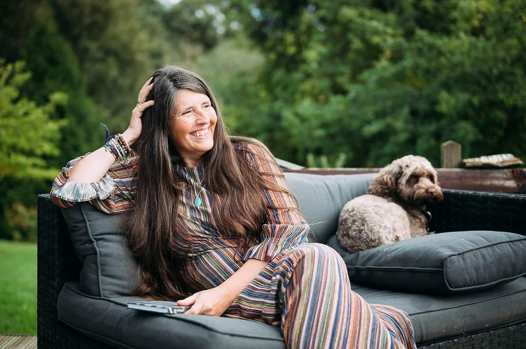 A woman with long brown hair, wearing a colorful striped dress, sitting on a gray outdoor sofa with a remote in her hand, smiling and resting her head on her hand. A small curly-haired dog is lying next to her on the sofa, with a background of green trees and foliage.