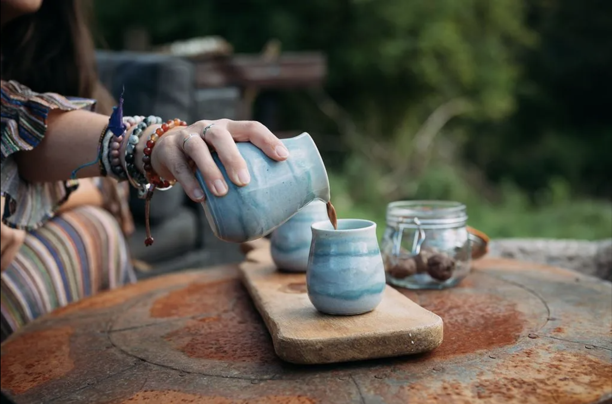 A person pours a hot cacao drink into handmade ceramic mugs on a rusted metal table outdoors.