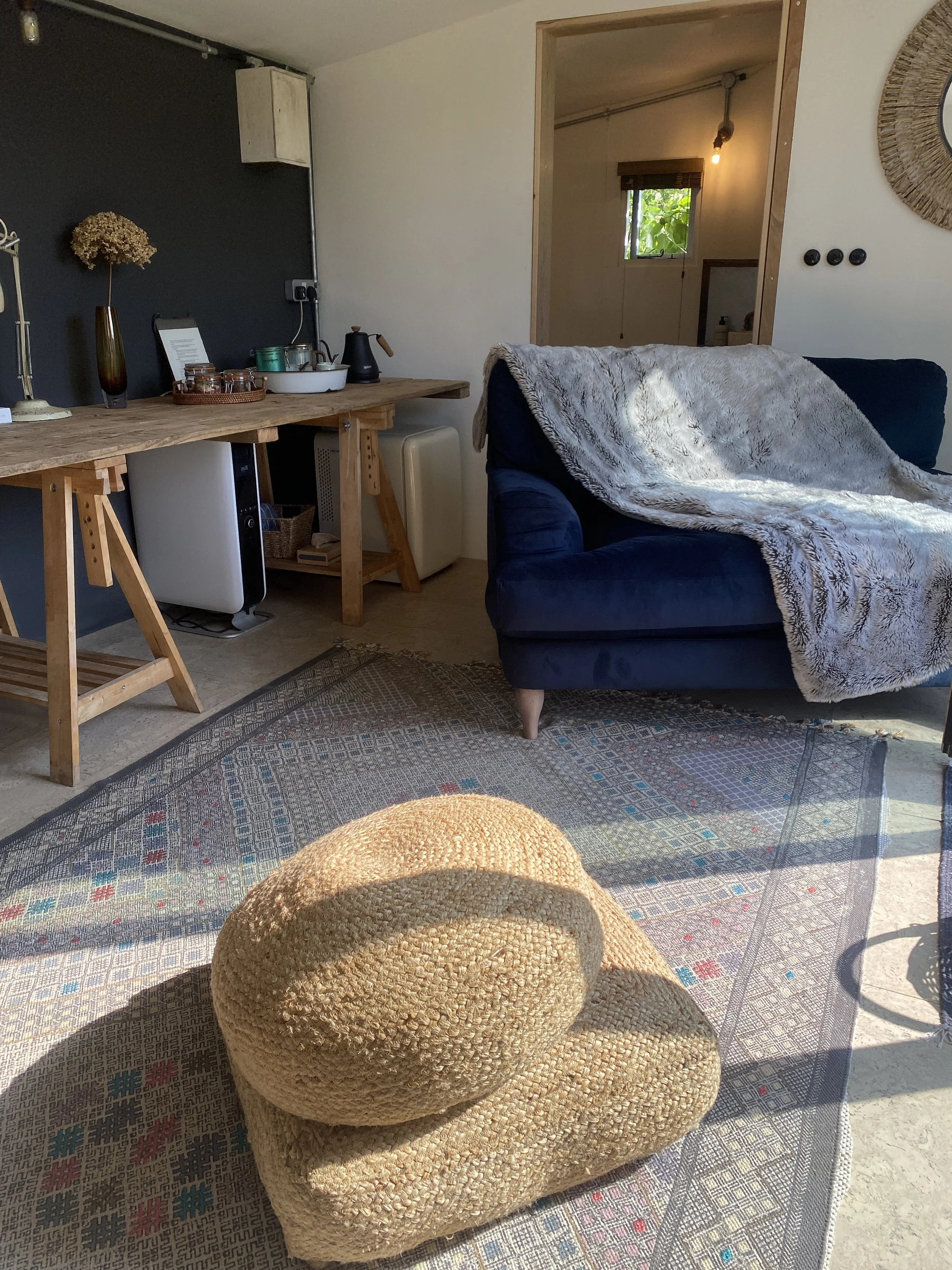 A cozy living room with a blue sofa covered by a fluffy blanket, a woven armchair, a wooden table with decorative items, and a rug on the floor. There is a doorway with a view to another room with a window and hanging light.