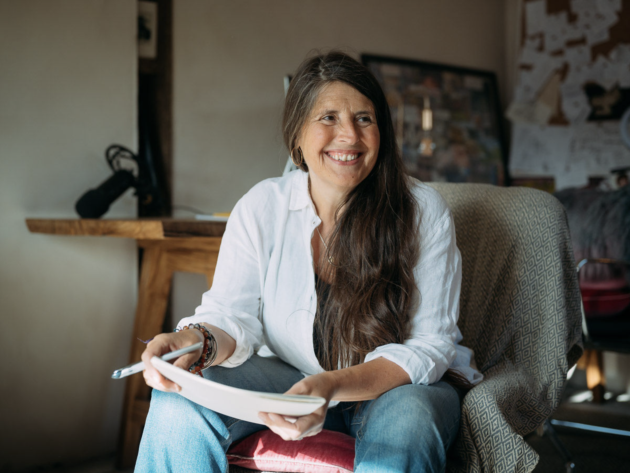 A woman with long brown hair, wearing a white shirt and blue jeans, is sitting in a chair indoors, smiling, holding a pen and a notebook.