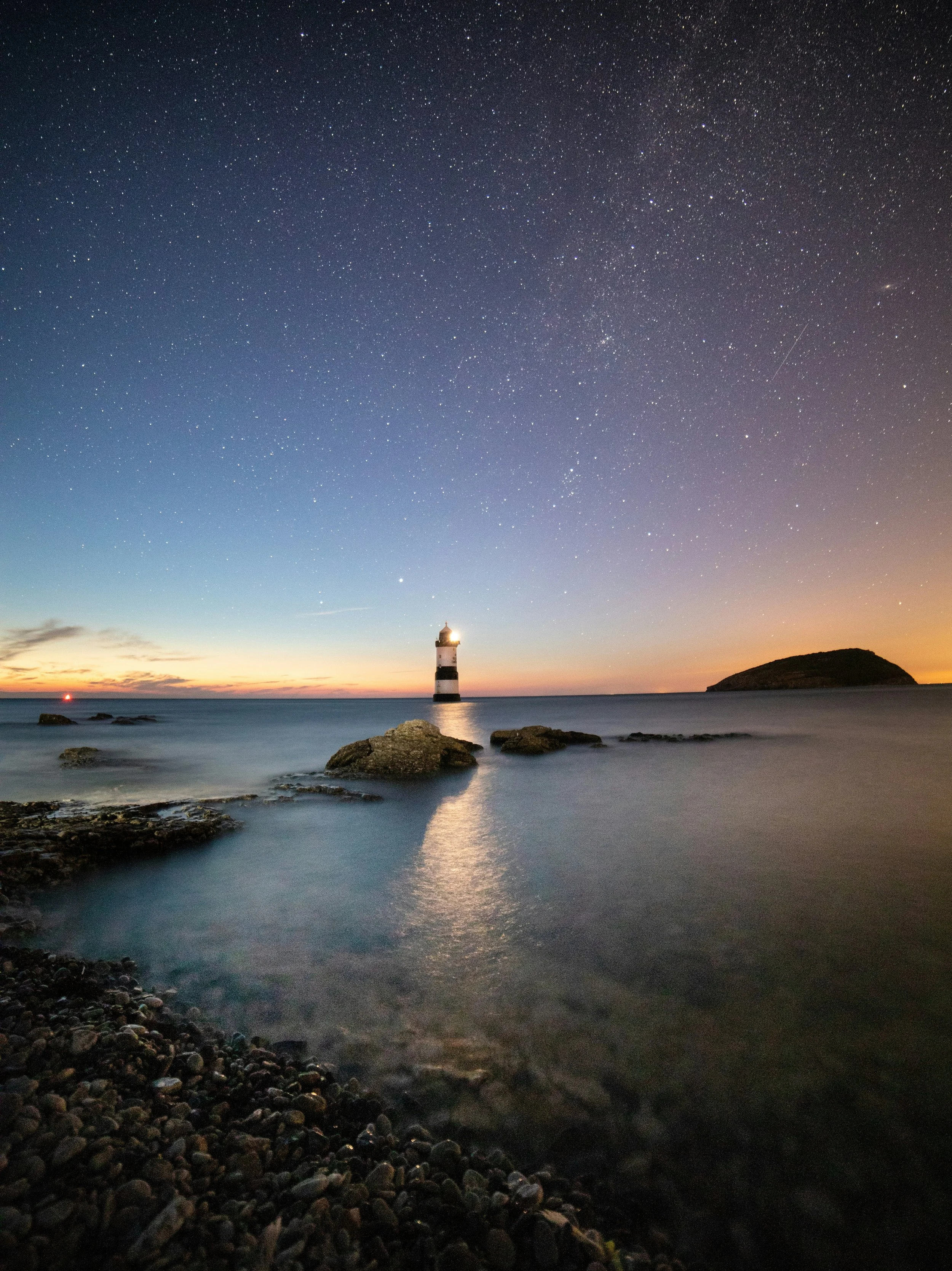 Starry night sky over a lighthouse near the ocean with rocks and a small island.