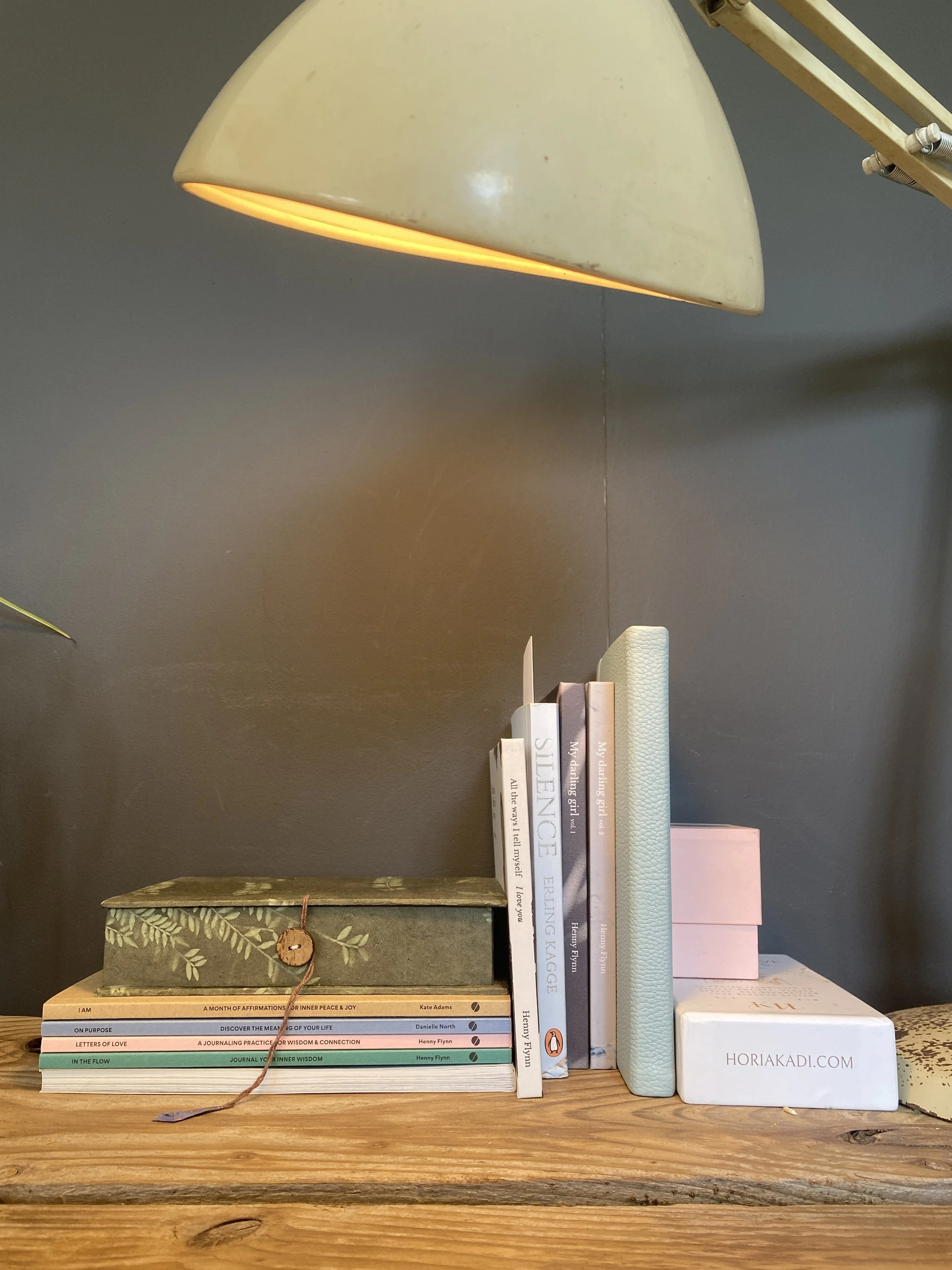 Books, a pink box, and a white box with the text 'Hori Kadi' on a wooden surface under a yellow desk lamp against a dark wall.