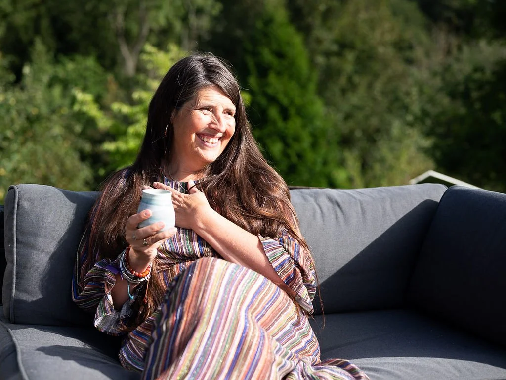 A woman with long brown hair sitting on an outdoor gray couch, smiling, holding a white mug, wearing a colorful striped dress, with greenery in the background.
