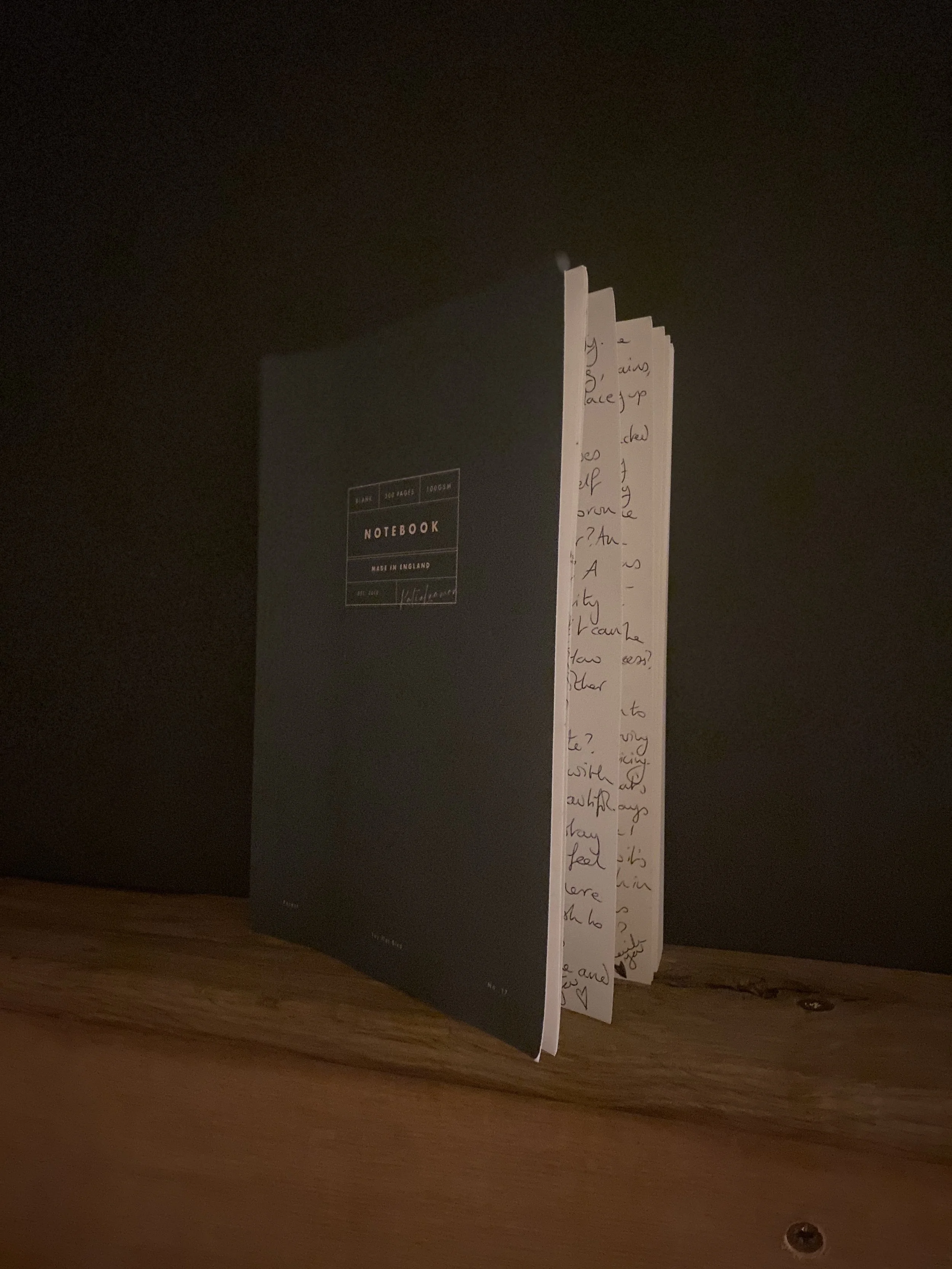 A dark green notebook with handwritten pages sticking out, placed on a wooden surface against a dark background.