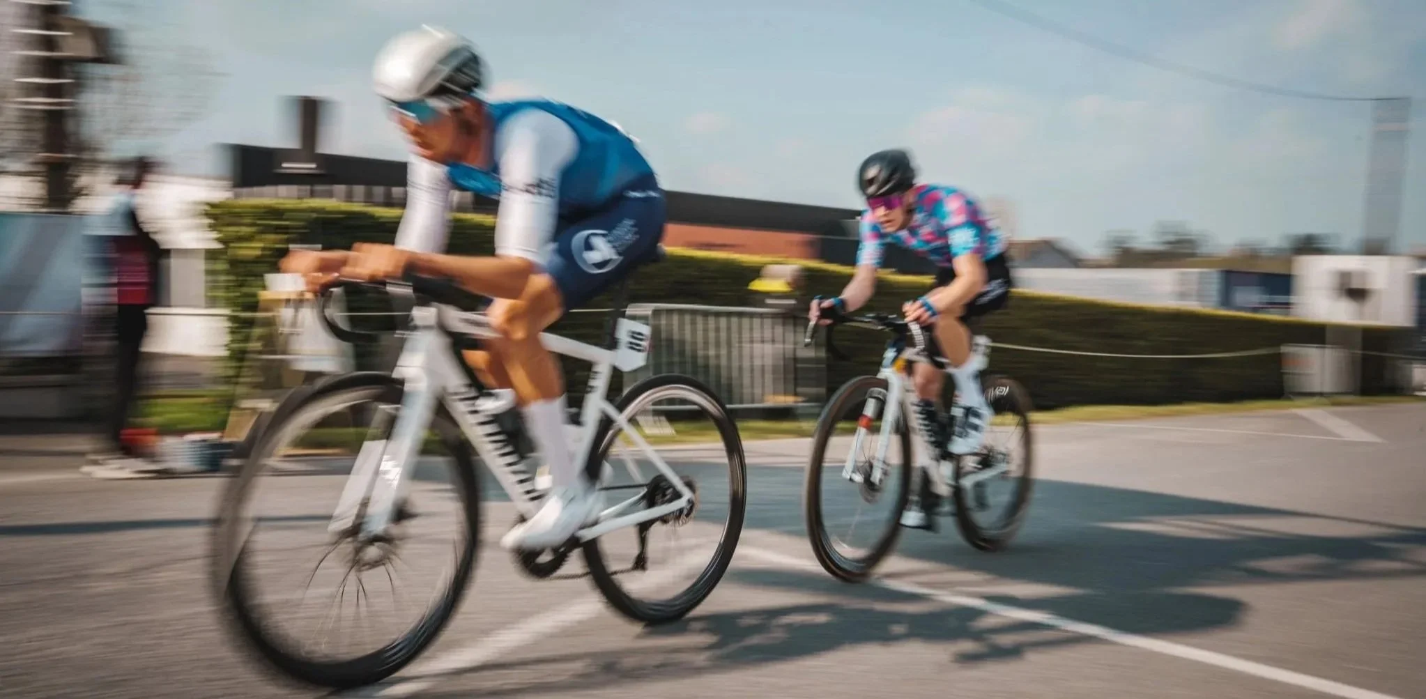 Deux cyclistes en course sur un circuit urbain, portant des casques et vêtements de sport, avec un public en arrière-plan.