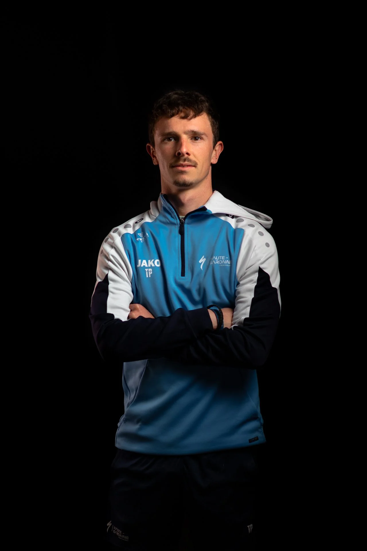 Un homme portant un uniforme de sport bleu et blanc, avec ses bras croisés, se tient contre un fond noir, avec une expression confiante.