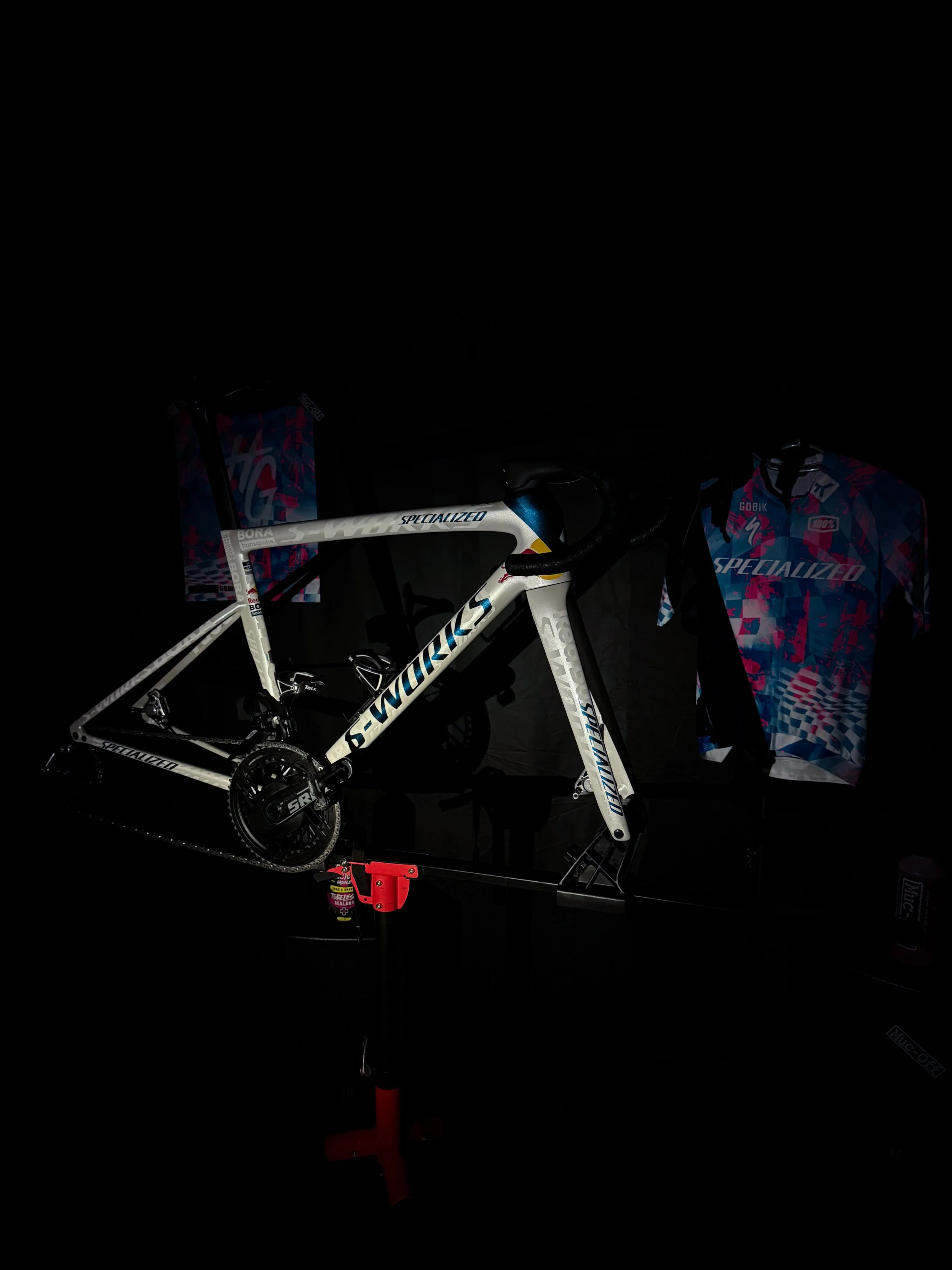 Vélo de route blanc suspendu sur un support. A l'arrière-plan, deux sacs ou tentes colorés avec des motifs bleus, roses et rouges.