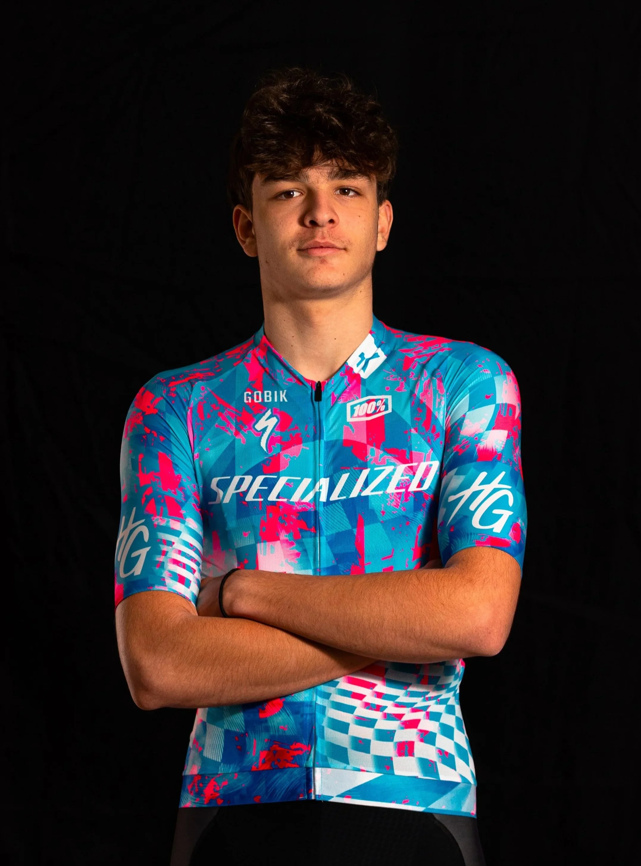 Portrait d'un jeune homme portant un maillot de cyclisme coloré avec des motifs en bleu, rose et blanc, portant un bras croisé, contre un fond noir.