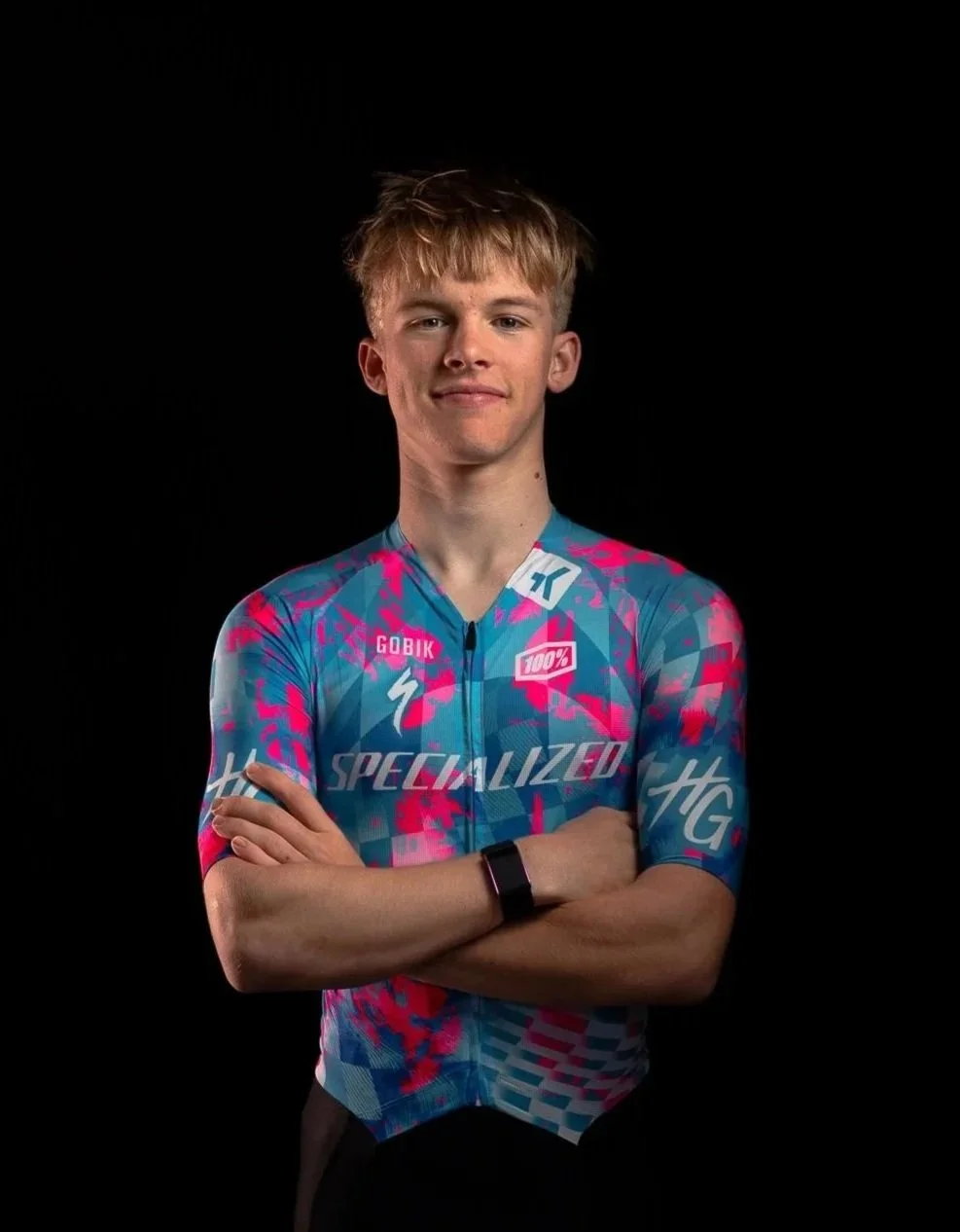 Un jeune homme debout avec des bras croisés, portant un maillot de cyclisme coloré avec des logos.