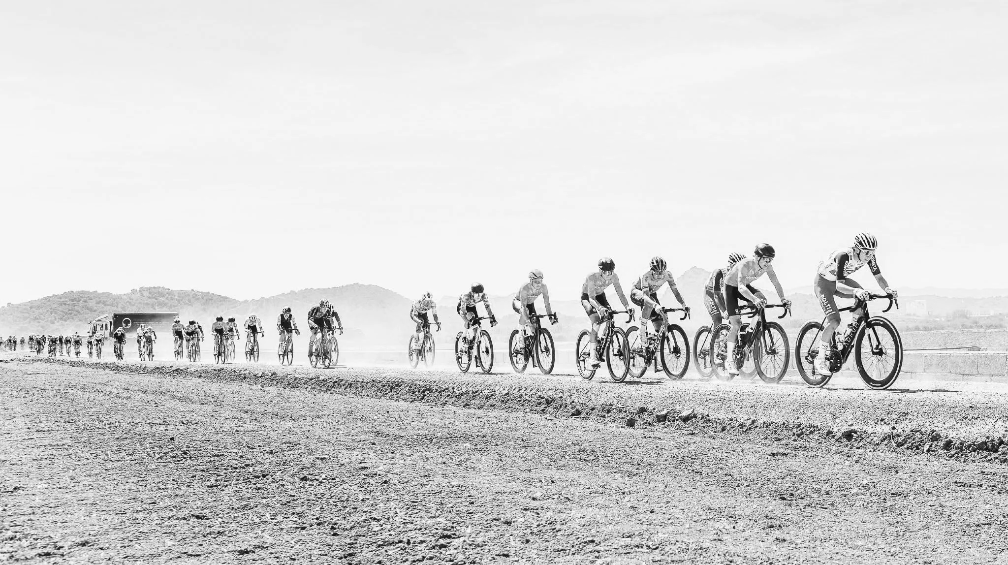 Groupe de cyclistes participant à une course sur un terrain désertique avec des montagnes en arrière-plan, en noir et blanc.